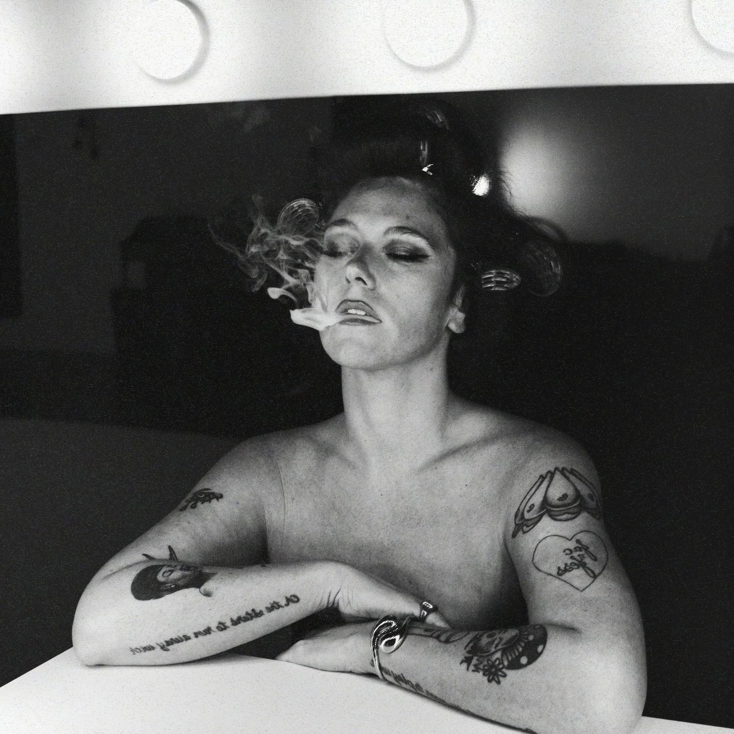 A black and white photograph of Suzanne Sheer with her eyes closed, surrounded by various tattoos on her body and accompanied by butterflies and handwritten text.