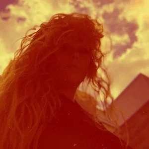 Suzanne Sheer with with long, curly hair standing outdoors at sunset, with a building in the background.