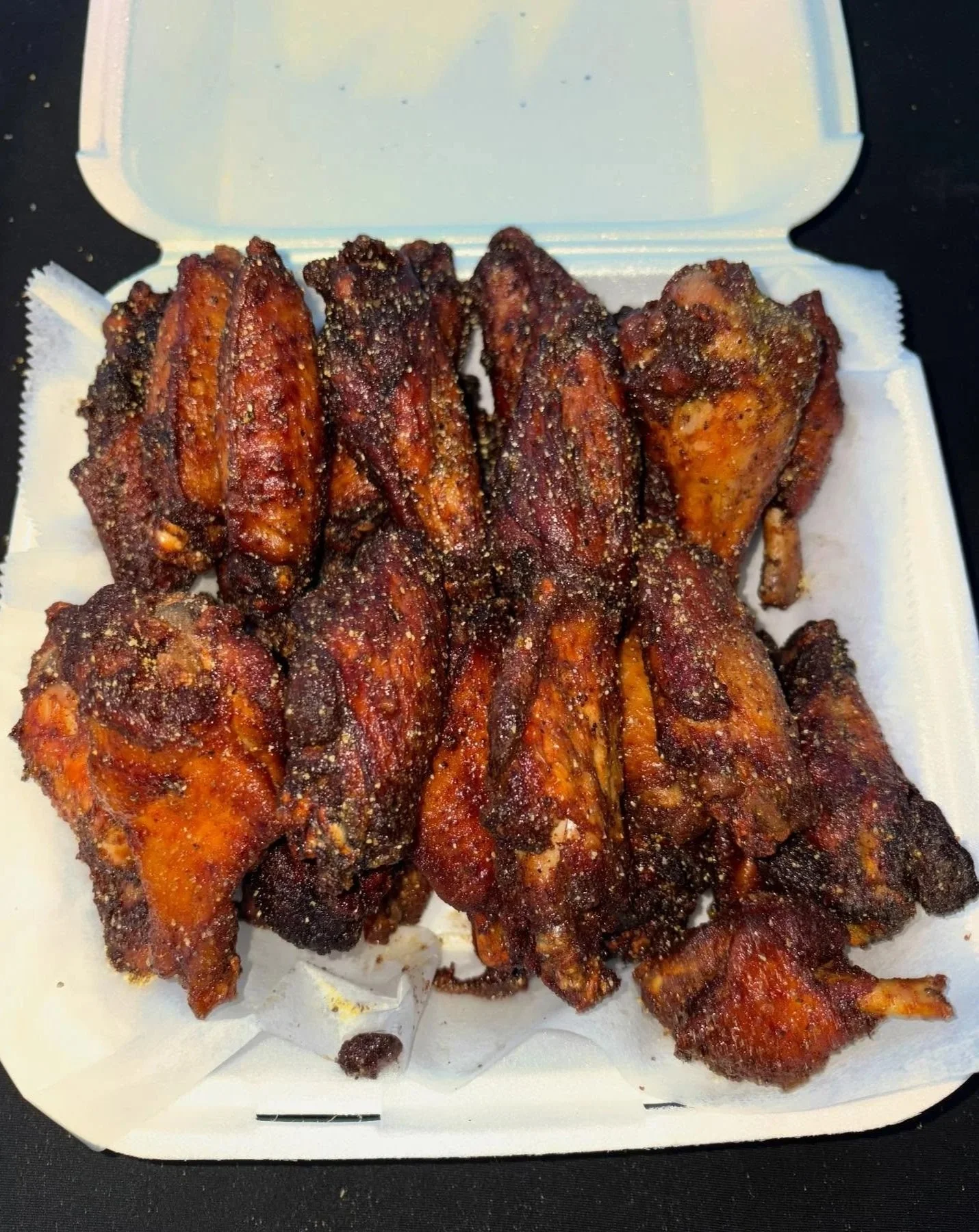 Close-up of a white takeout container filled with spicy baked chicken wings garnished with black pepper.