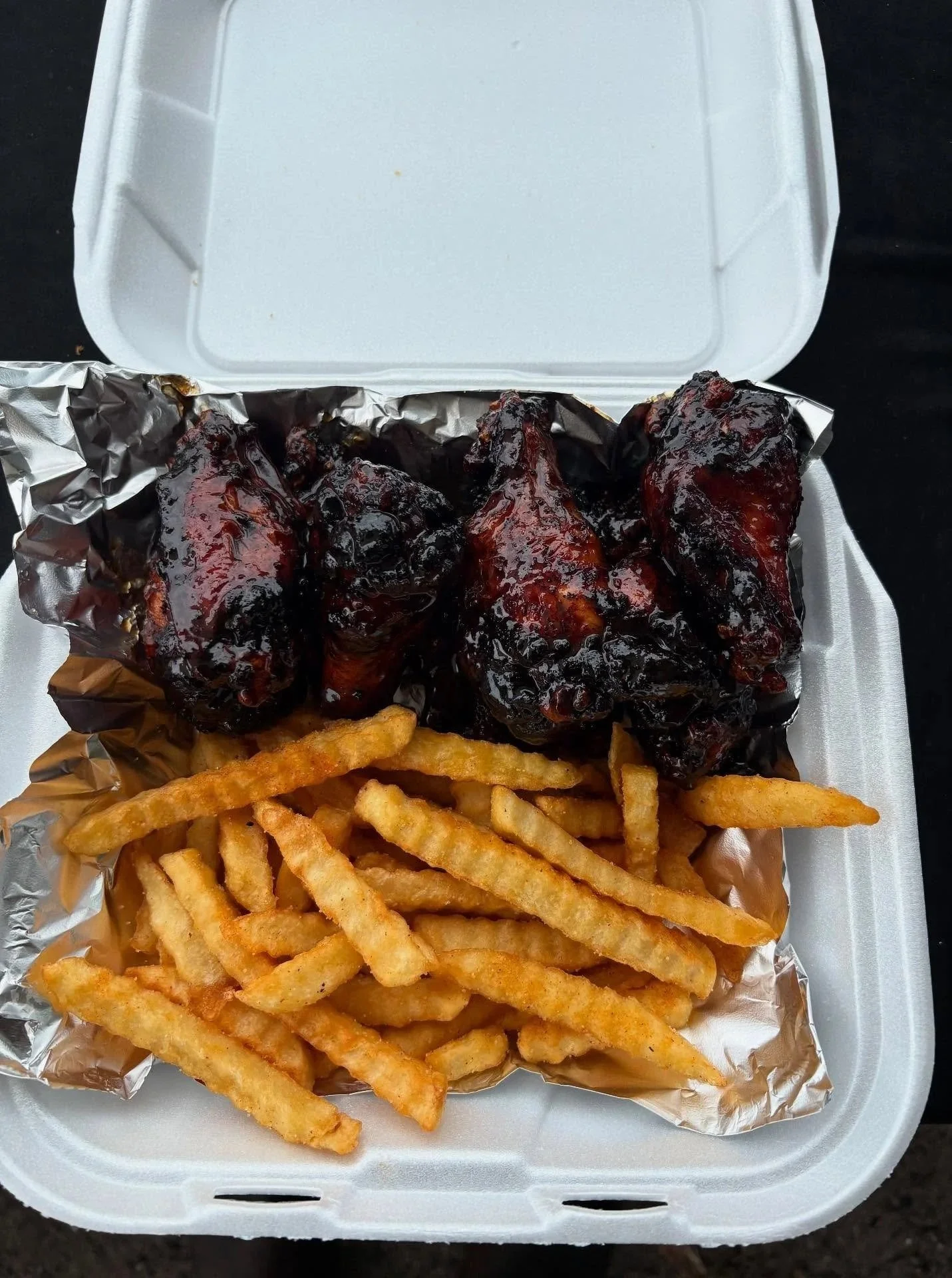 To-go container with French fries and barbeque chicken drumsticks covered in sauce.