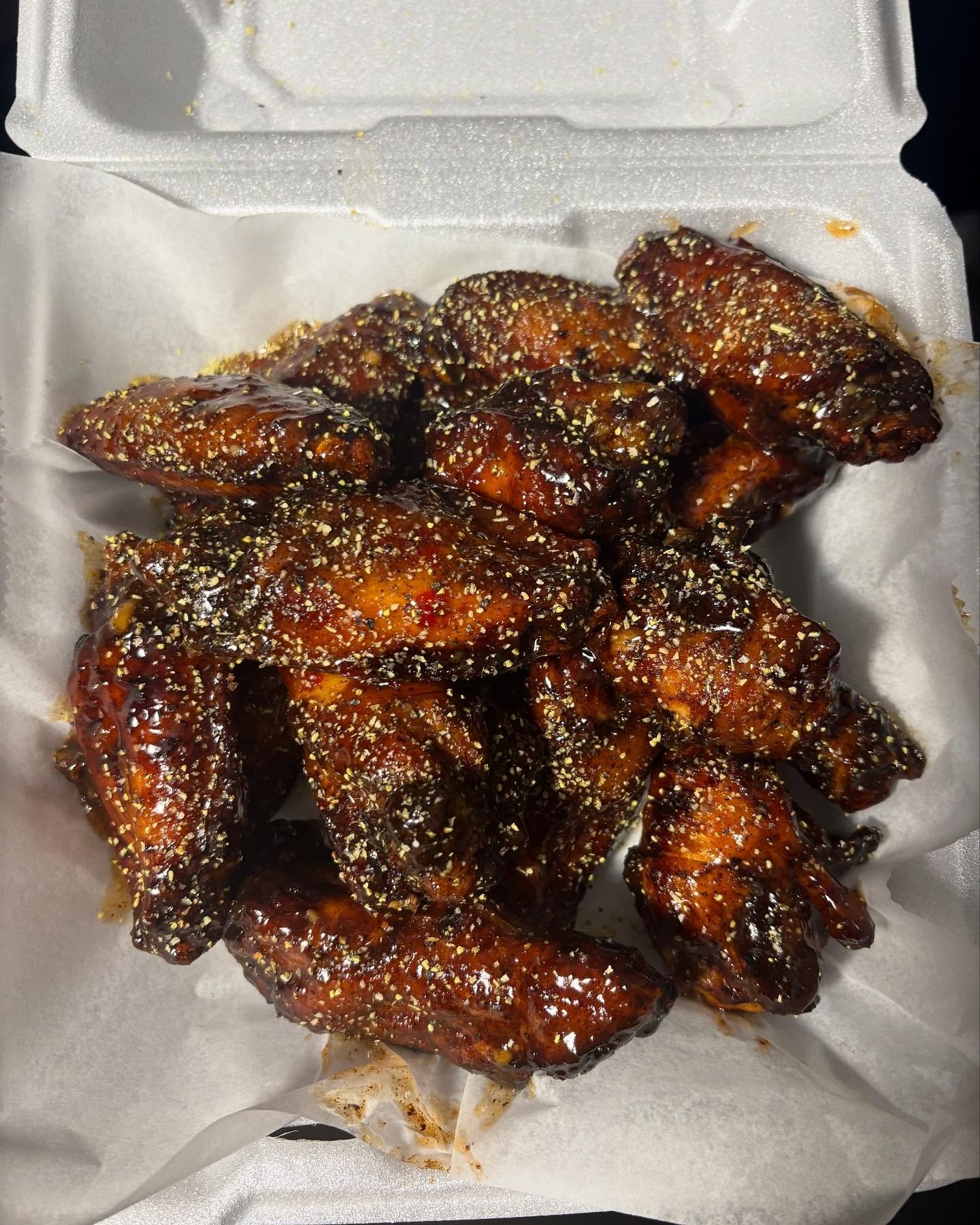 Close-up of glazed chicken wings topped with sesame seeds in a foam food container lined with parchment paper.