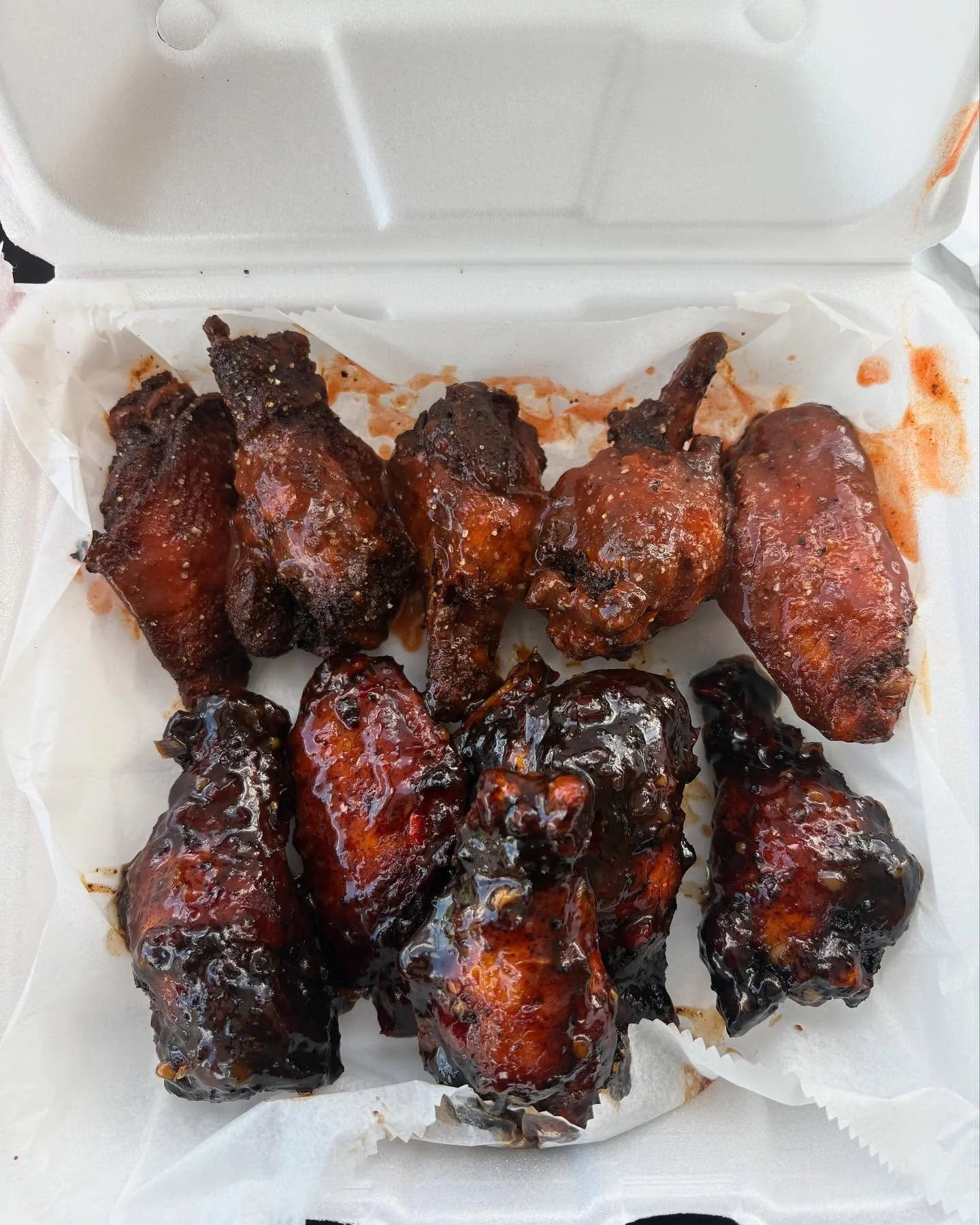Box of saucy, grilled chicken wings with charred edges, covered in barbecue sauce, on white parchment paper