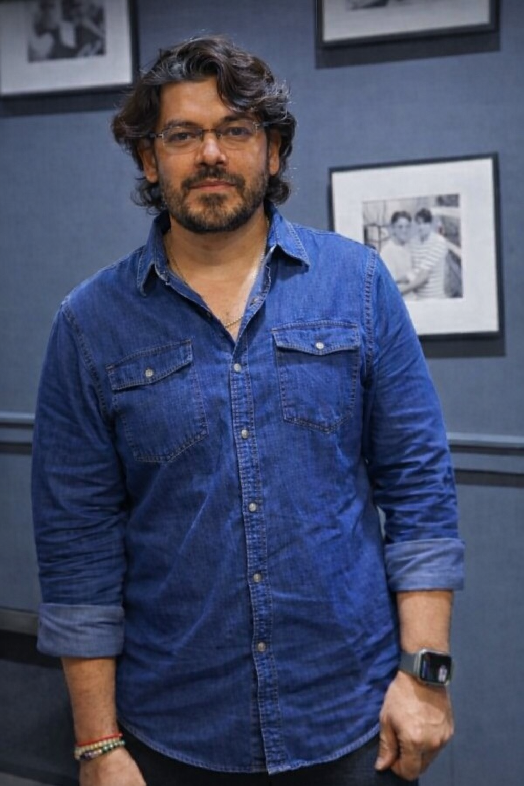 A man with glasses and curly hair, wearing a long-sleeved denim shirt, standing in front of framed black-and-white photographs on a gray wall.