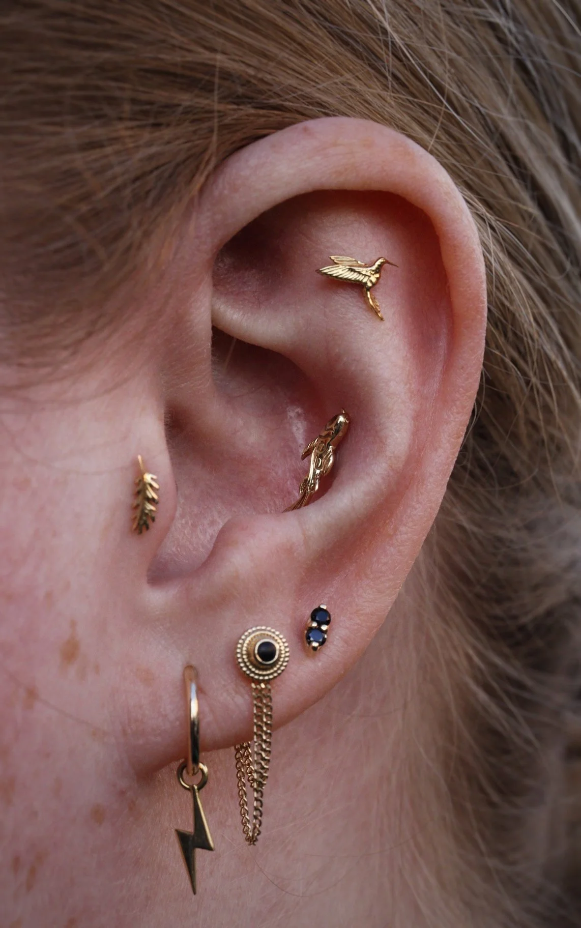 Varios aretes dorados en la oreja, incluyendo uno en forma de colibrí y otros con diferentes diseños.