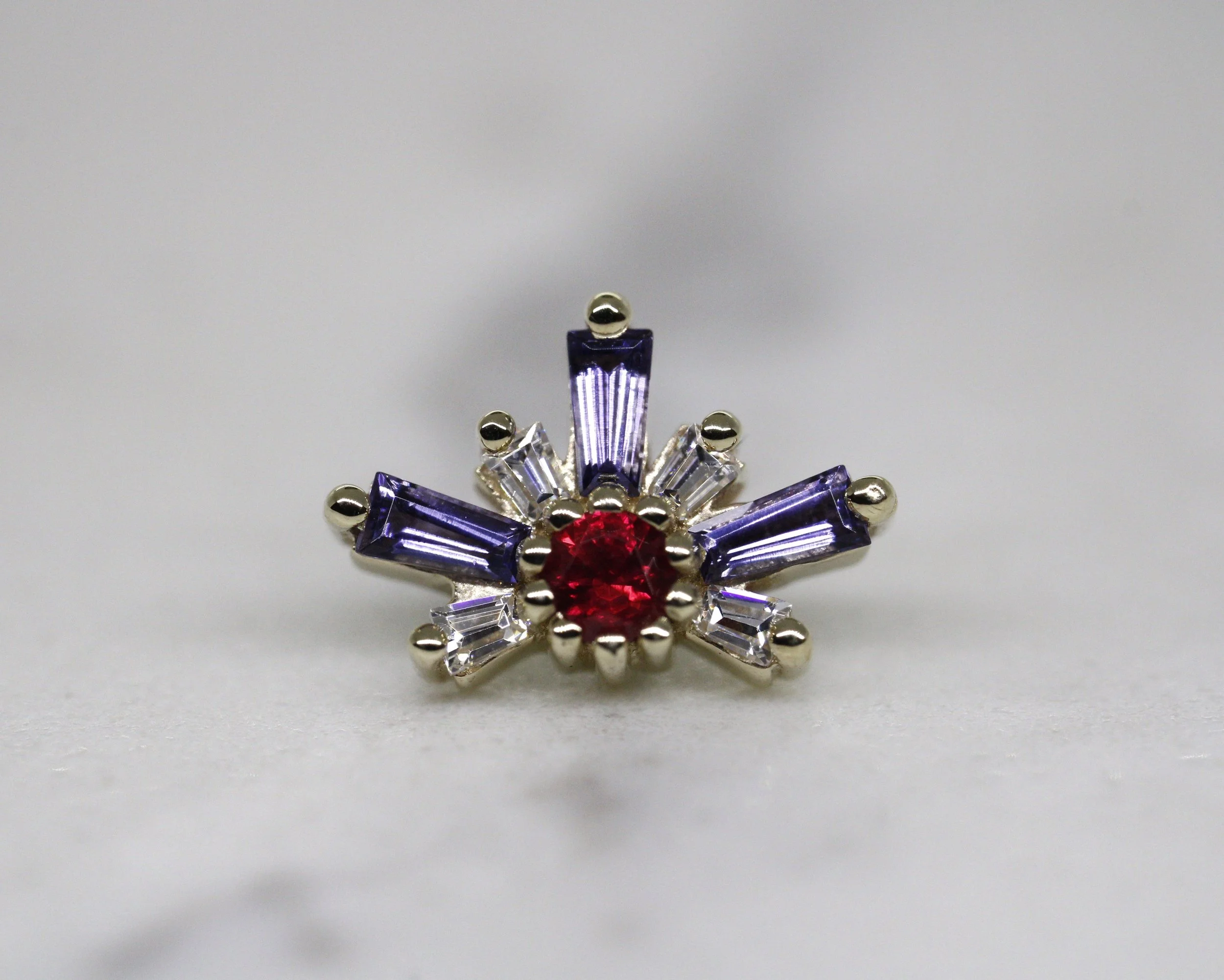 Joya de forma floral con piedras moradas, rojas y plateadas sobre fondo gris claro.