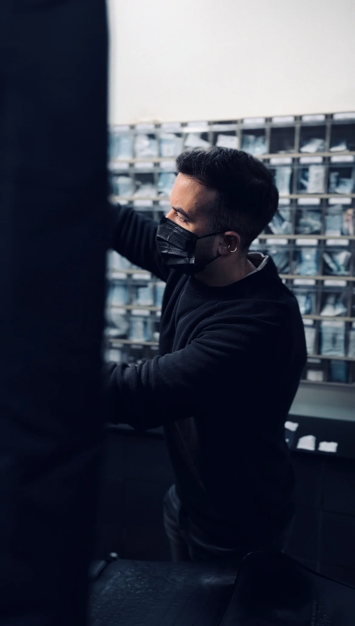 Joven con barba y peinado moderno usando mascarilla negra, en un entorno con estanterías llenas de productos, posiblemente en una tienda o almacén.