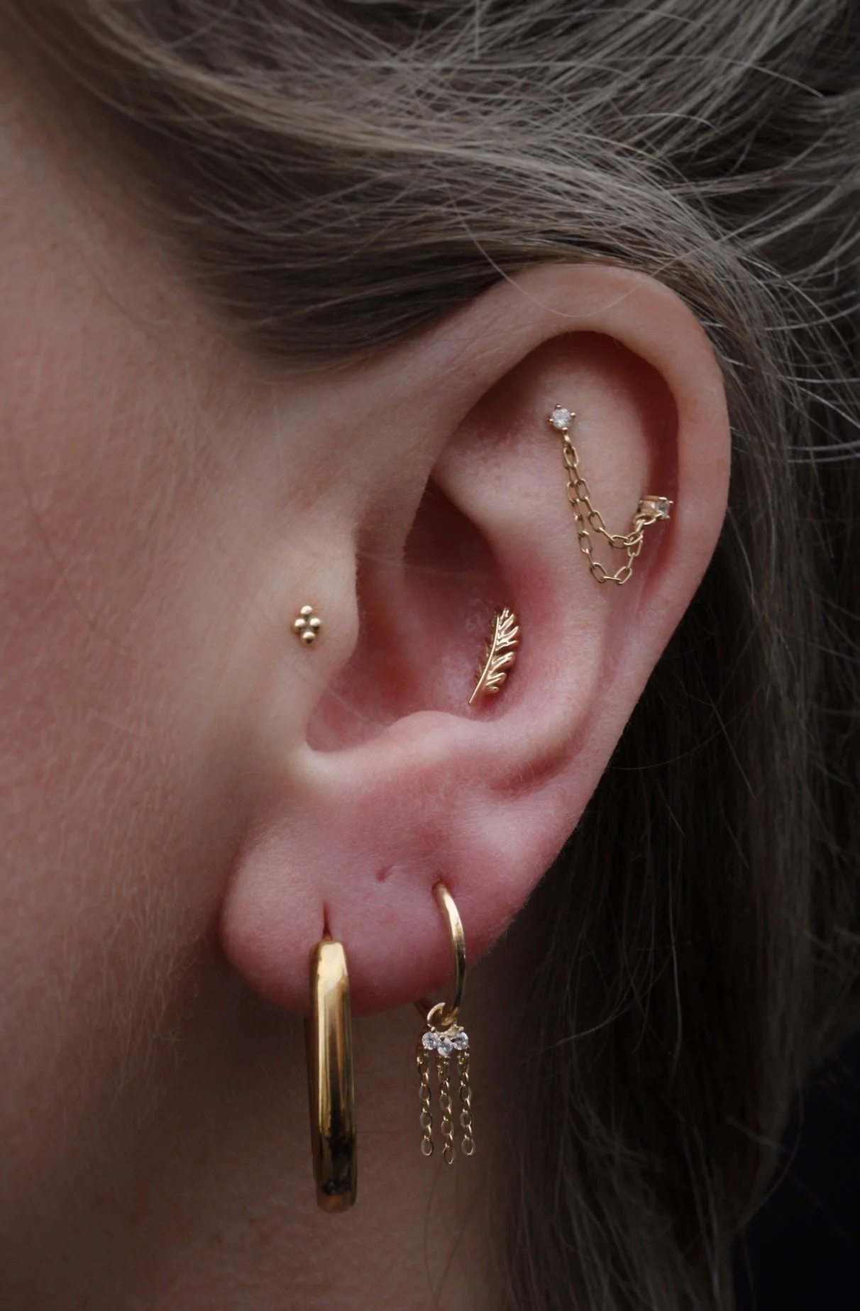 Varios pendientes de oro, incluyendo uno con cadena, uno en forma de hoja, uno con pequeña piedra y otros. Un peinado en cabello rubio y parte de rostro visible.