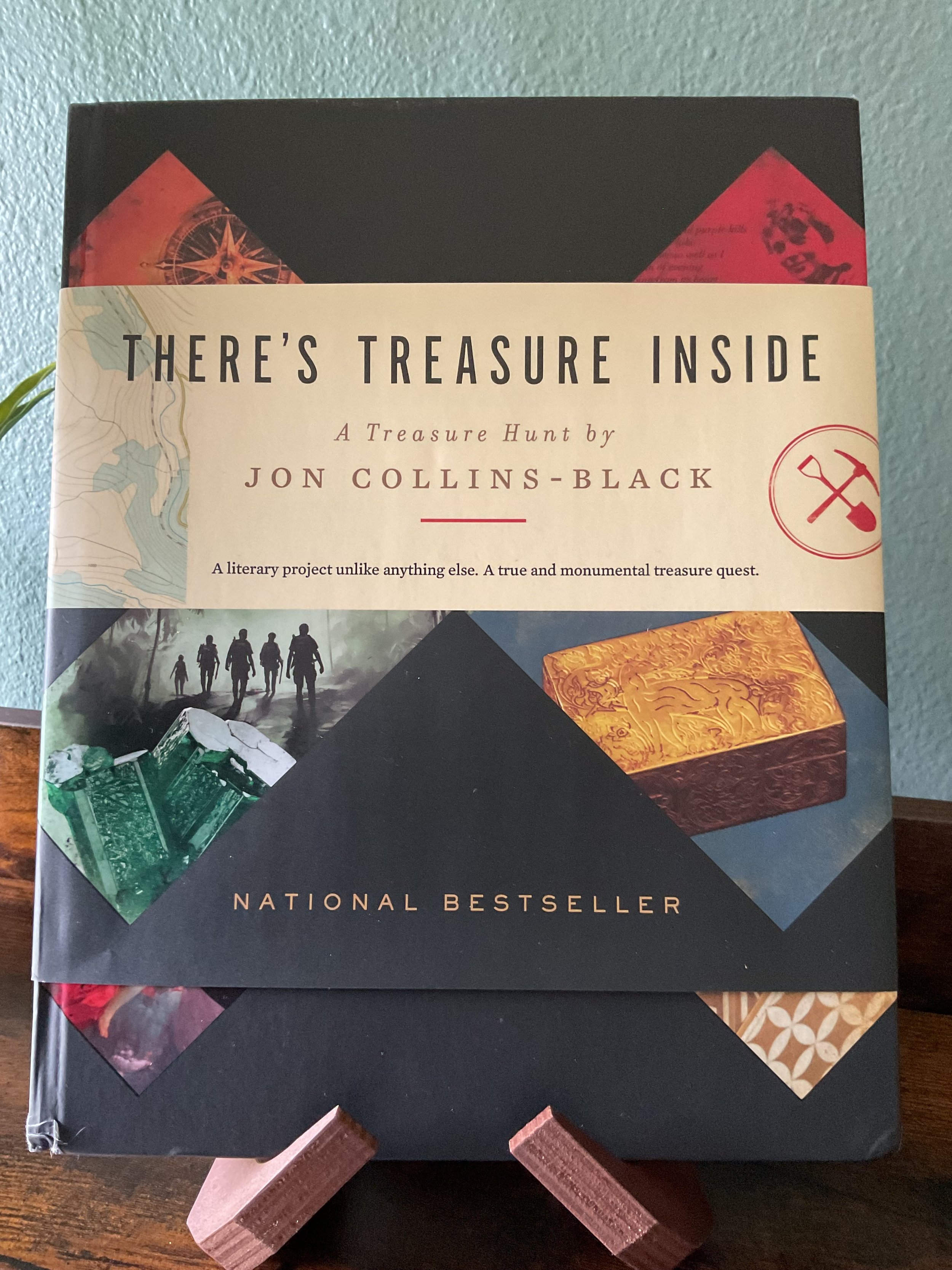Book titled 'There's Treasure Inside' by Jon Collins-Black with a compass, map, and treasure chest illustrations on the cover, resting on a black surface with a wooden table background.