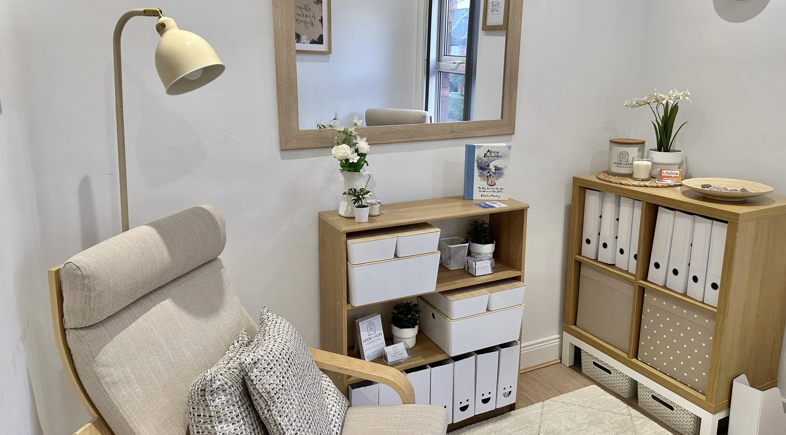 A cozy interior corner with a beige armchair, a wooden shelf with storage boxes and plants, a wall mirror, and decorative items including a flower arrangement, framed digital illustration, and a shelf with binders and decor items.