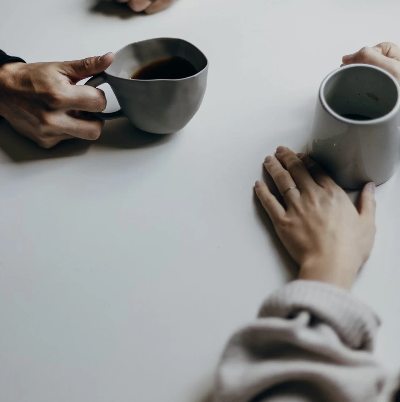 Two people are holding ceramic cups with dark beverages, sitting at a white table, one with a gold band on finger.