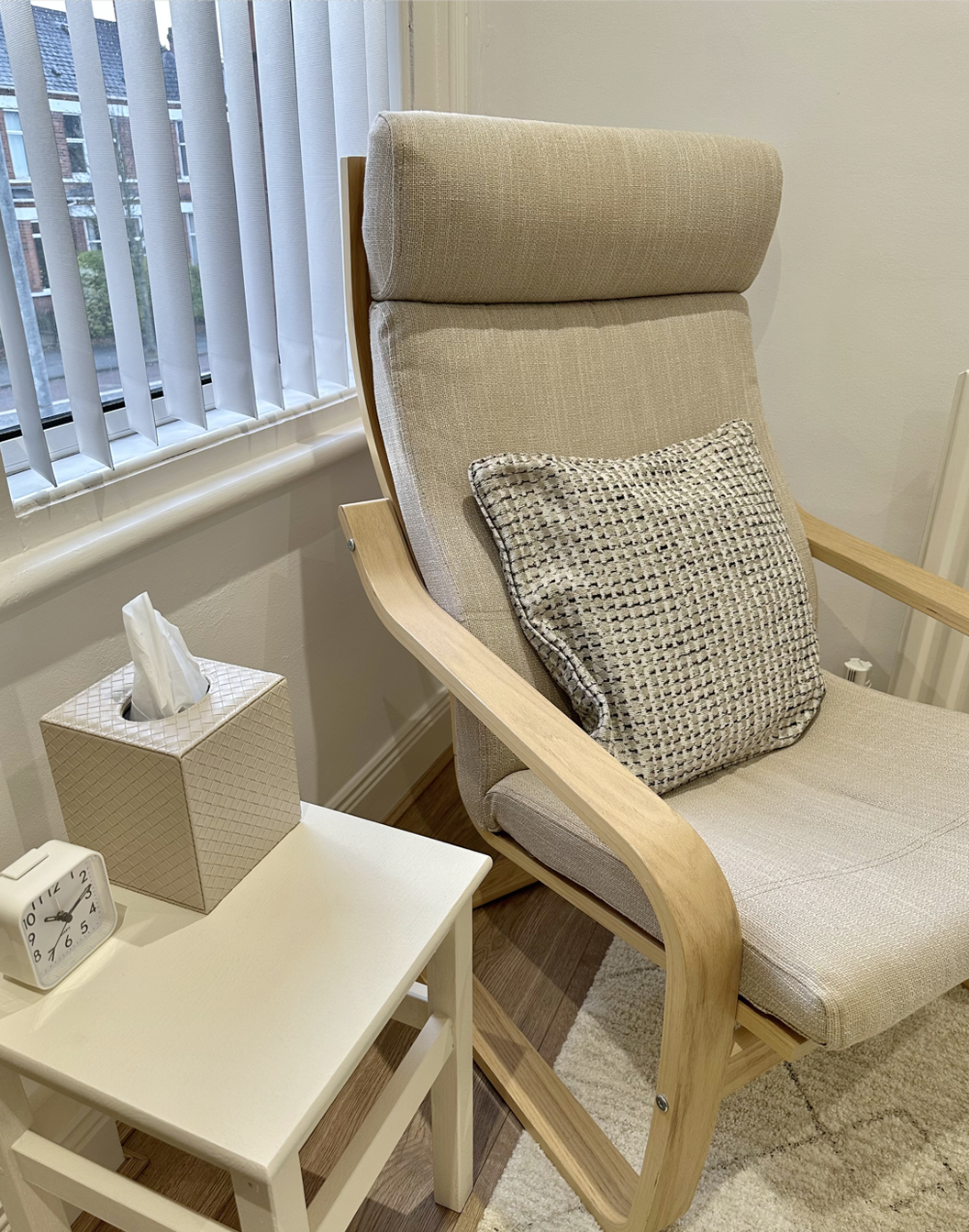 A beige upholstered armchair with a trapped pillow and light wooden frame in a room next to a white side table with a tissue box, an alarm clock, and a window with vertical blinds.