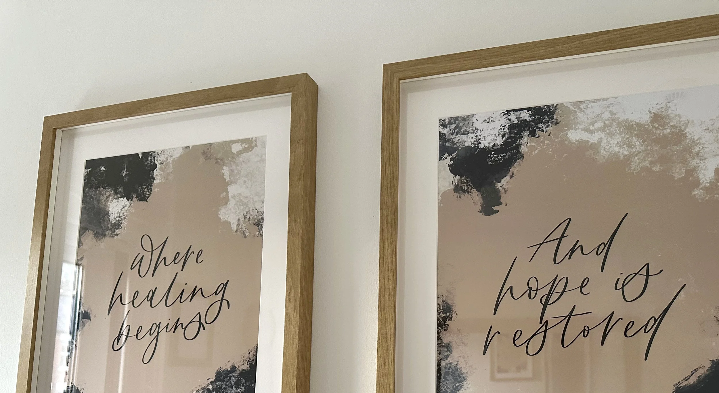 Close-up of two framed prints with abstract black, white, and beige backgrounds and cursive text about hope and healing hanging on a white wall.