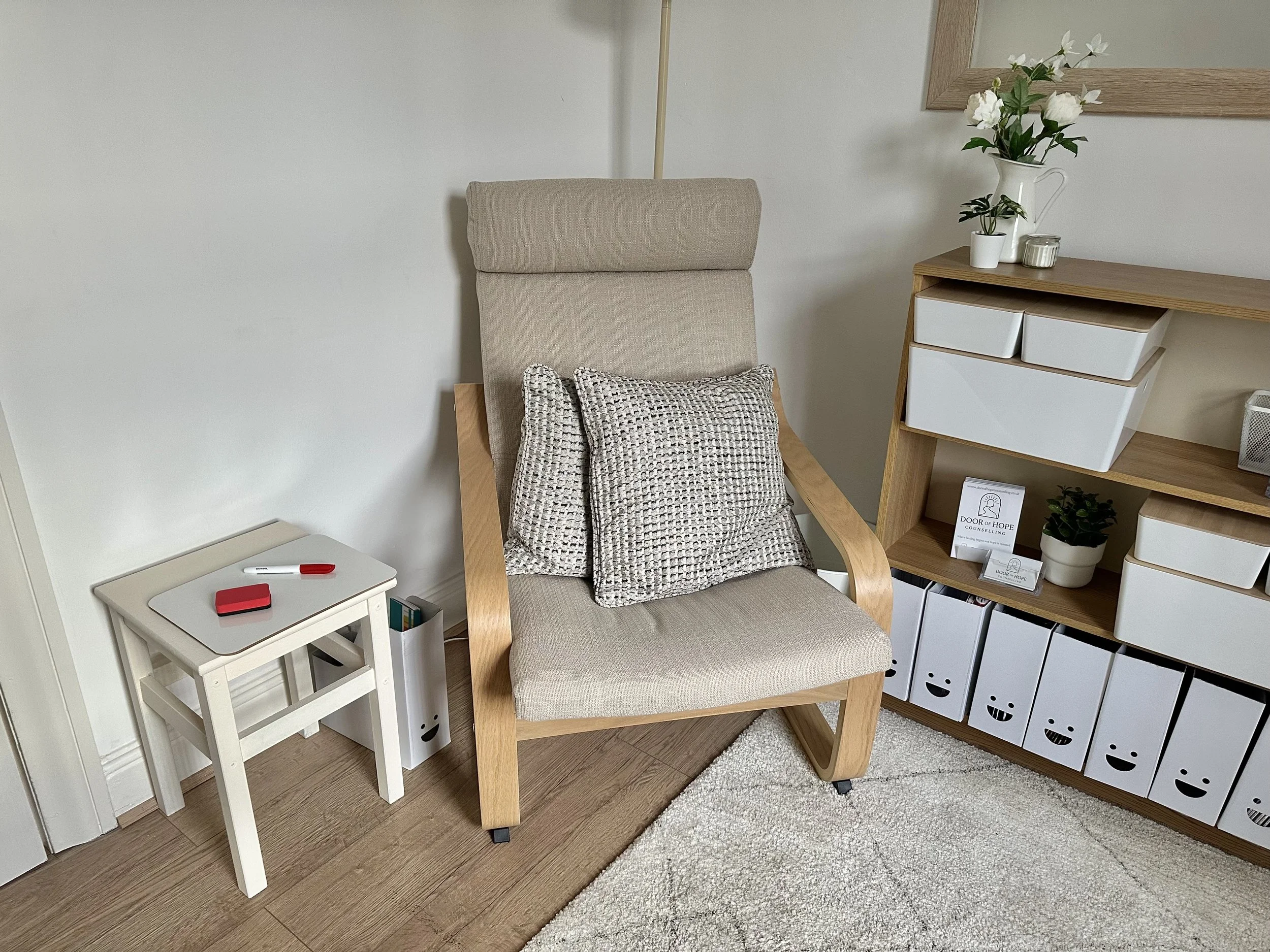 A beige upholstered armchair with a patterned pillow on it, next to a small white side table with a red marker and object on top, beside a storage unit with white drawers and a wooden top, decorated with a white pitcher with flowers and a small potted plant.