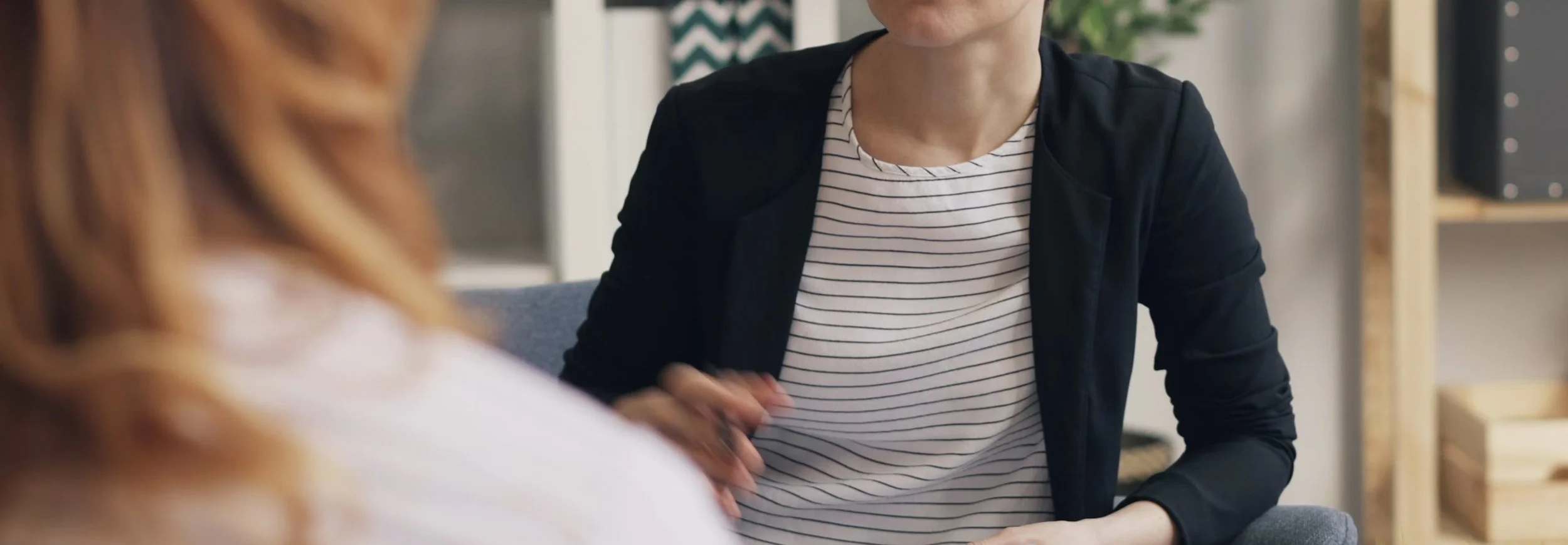 A person in a black jacket and striped shirt shaking hands with a woman in a white shirt with blurred red hair in a modern office setting.