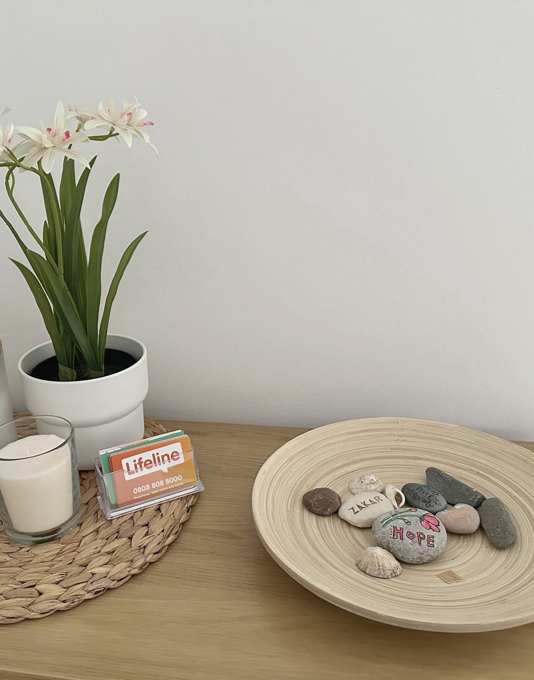 A wooden table with a potted white orchid, a lit candle, and a small stack of colorful Lifeline cards on a woven mat. A round wooden tray holds decorated and plain stones with words like 'HOPE' and 'ZAKAR'.
