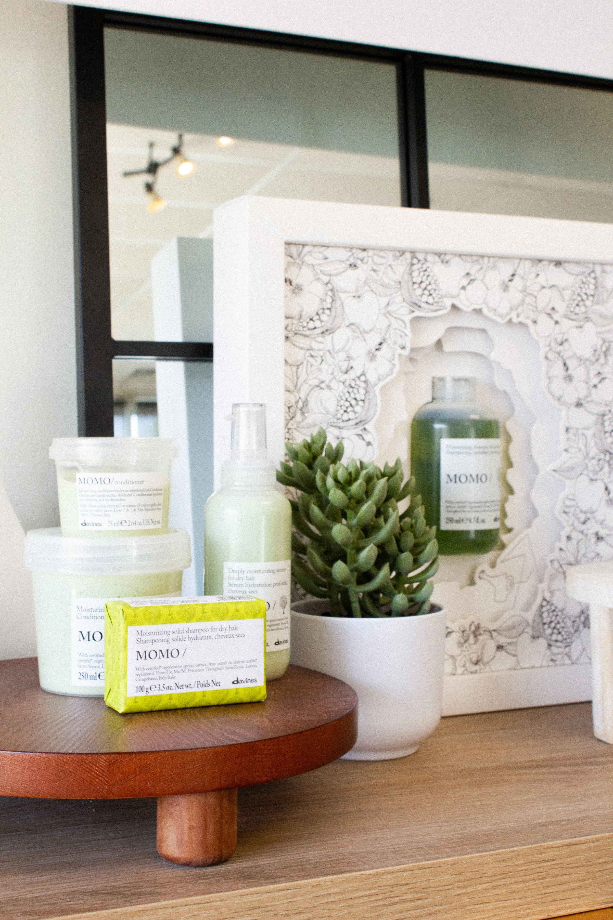 Display of Davines momo haircare products, including several tubes and bottles, with a potted succulent plant and a decorative ornate paper cutout in the background.