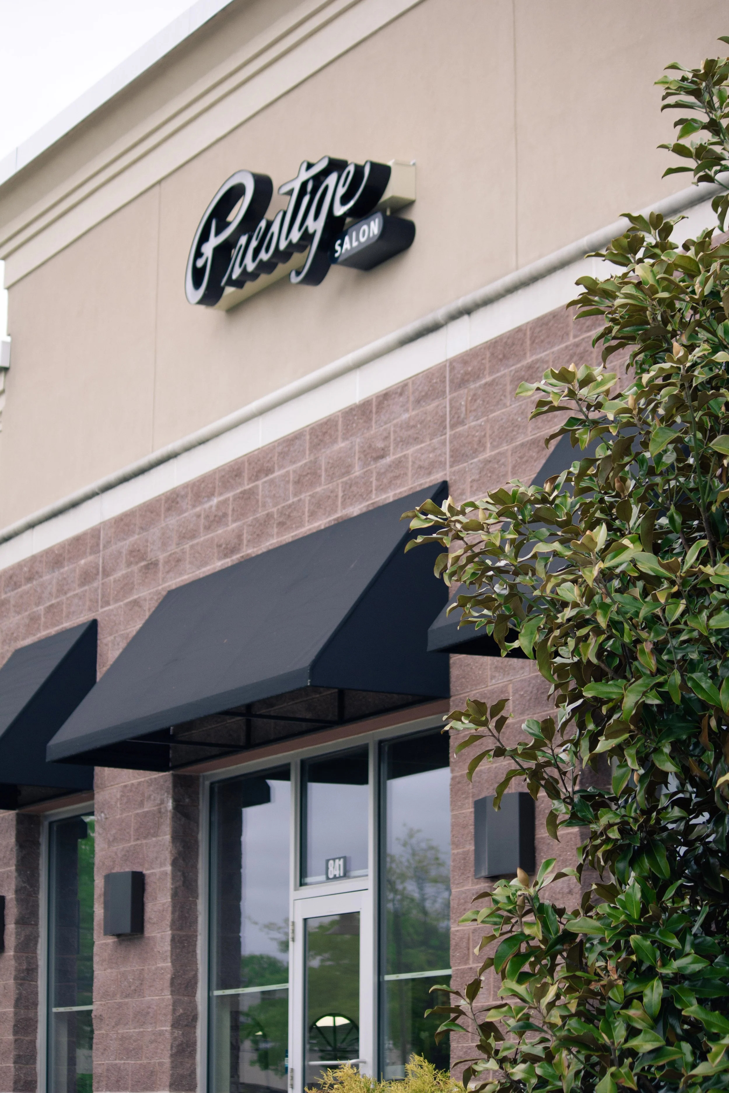 Exterior view of a building with a sign that reads "Prestige Salon". The building has brick and beige walls with black awnings over the windows and a leafy tree in the foreground.