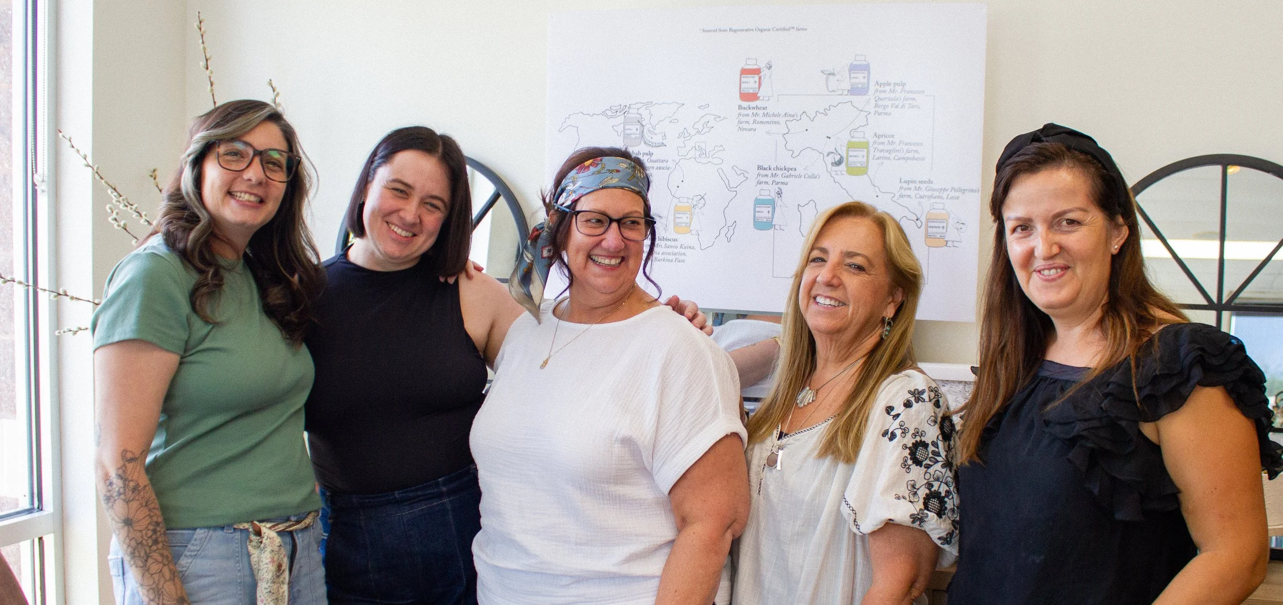 Five women standing together indoors, smiling, with a world map poster behind them.