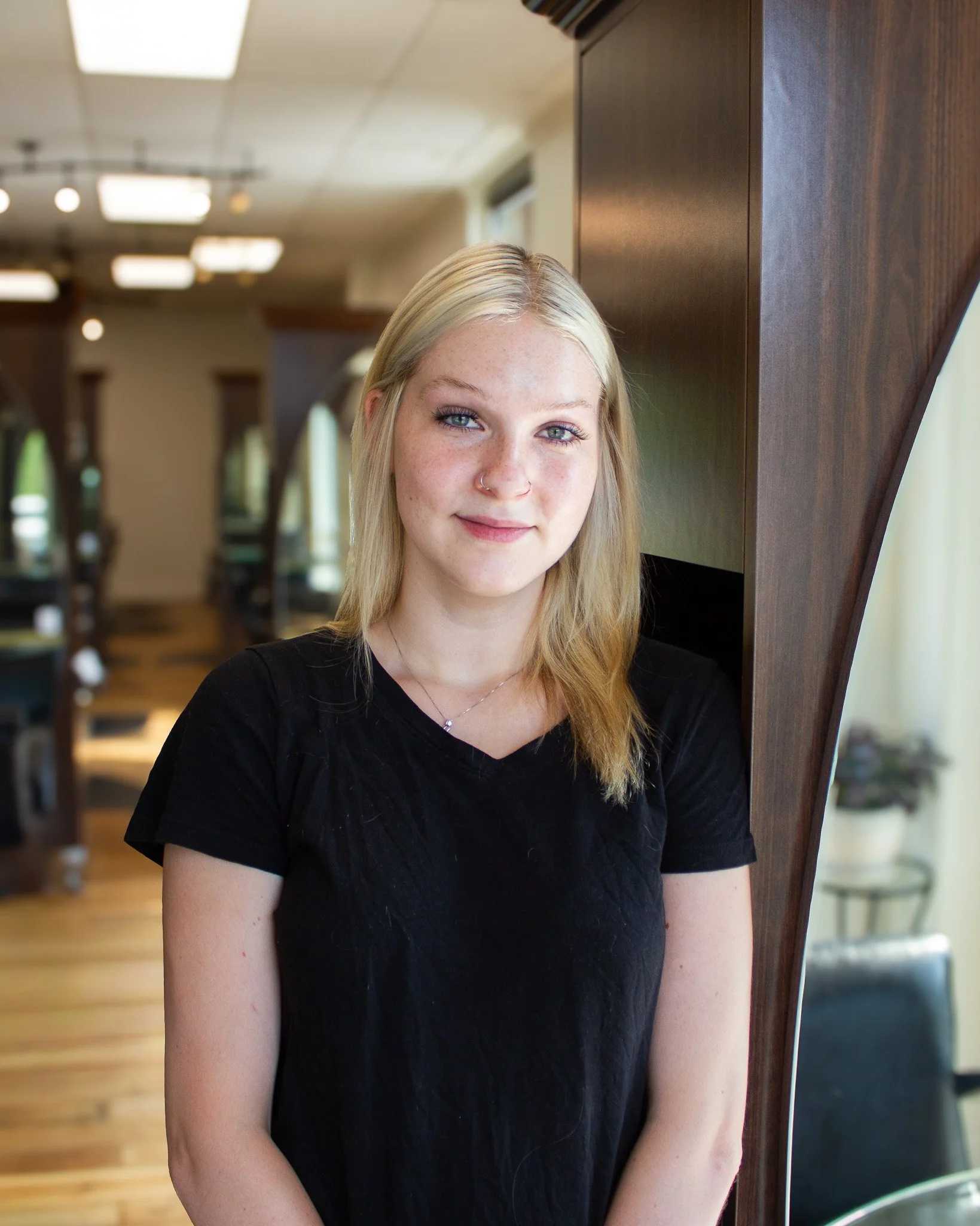 A young woman with long blonde hair, wearing a black shirt, standing indoors next to a wooden partition, with a blurred background of a room with ceiling lights and bookshelves.