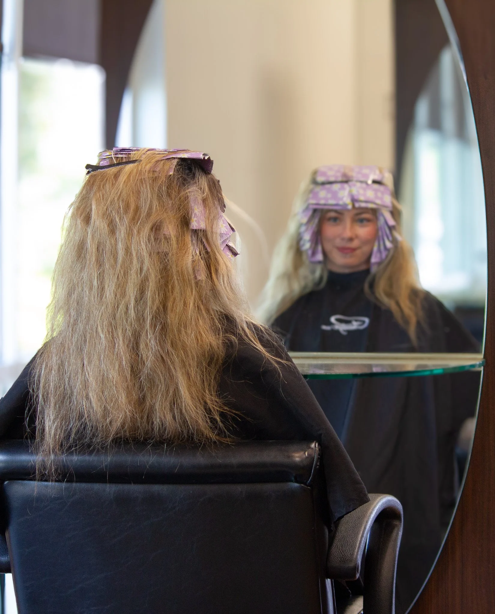 A woman with long blonde hair and purple curlers in her hair, looking at herself in the mirror, sitting in a salon chair.