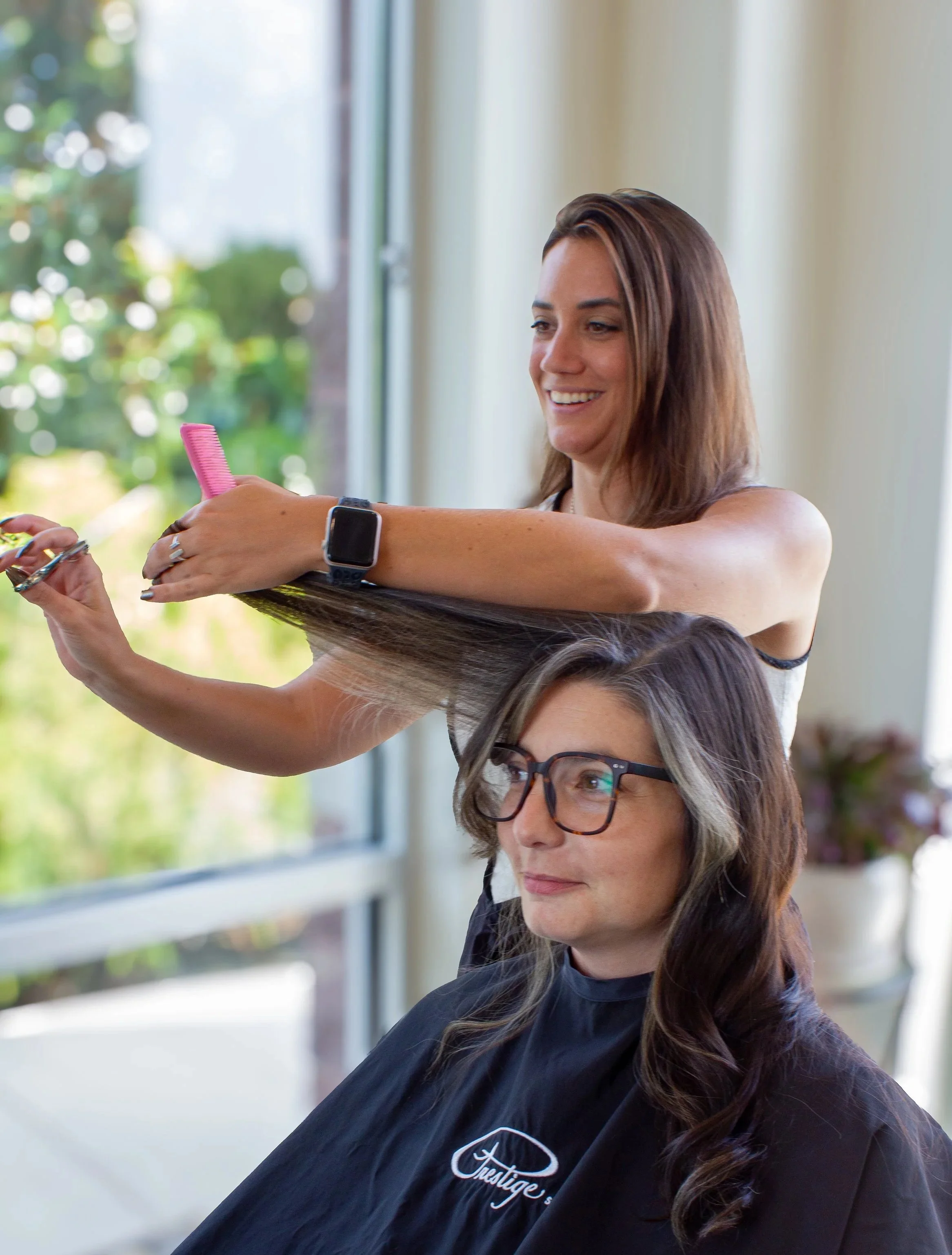 A woman with long brown hair wearing glasses and a black salon cape sits in a hair salon chair, while a hairstylist with brown hair and a smartwatch styles her hair using a pink comb. The background features a window and green foliage outside.