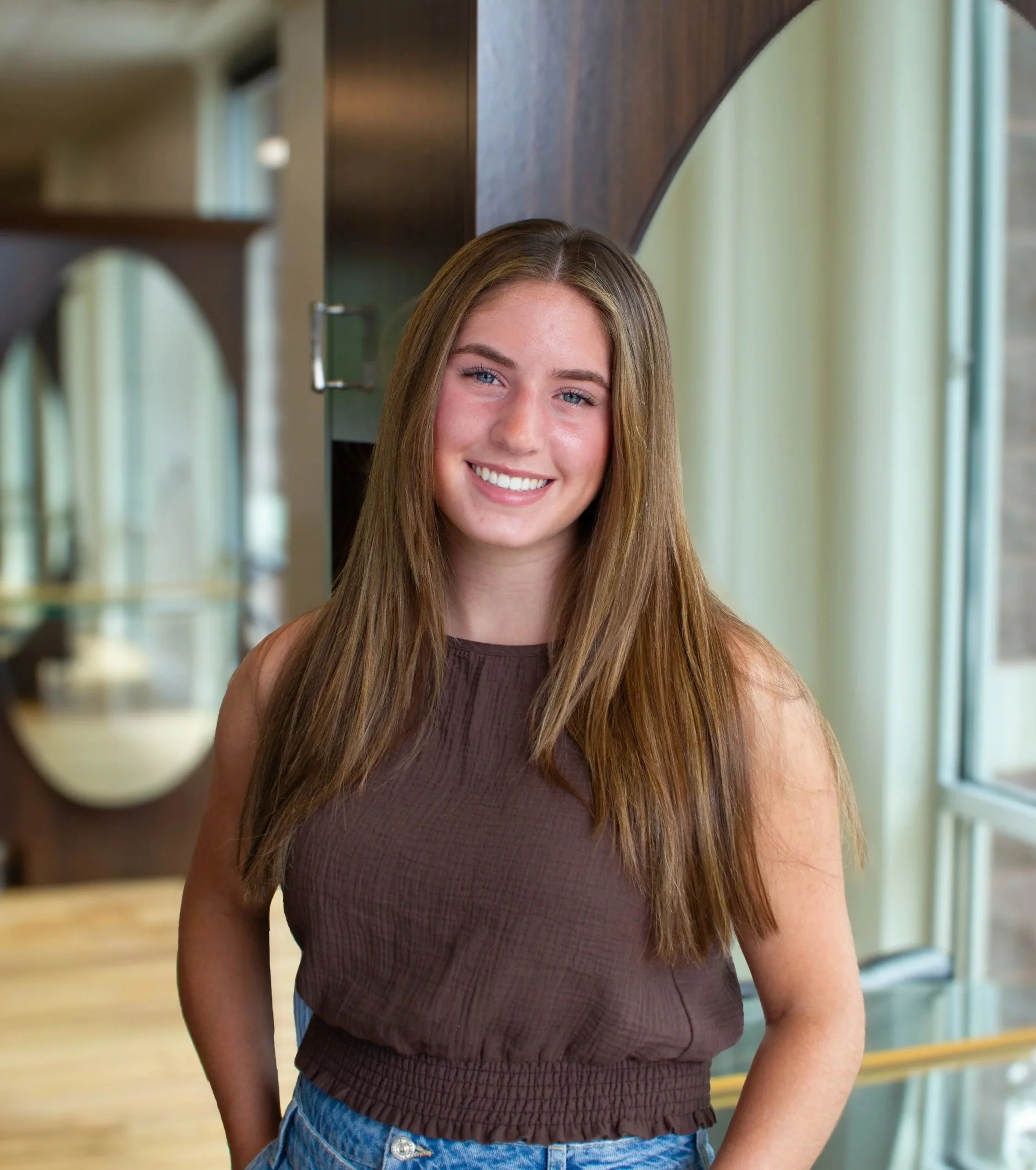 Young woman with long light brown hair and blue eyes smiling, standing indoors near a window and bookshelf, wearing a sleeveless brown top and jeans.