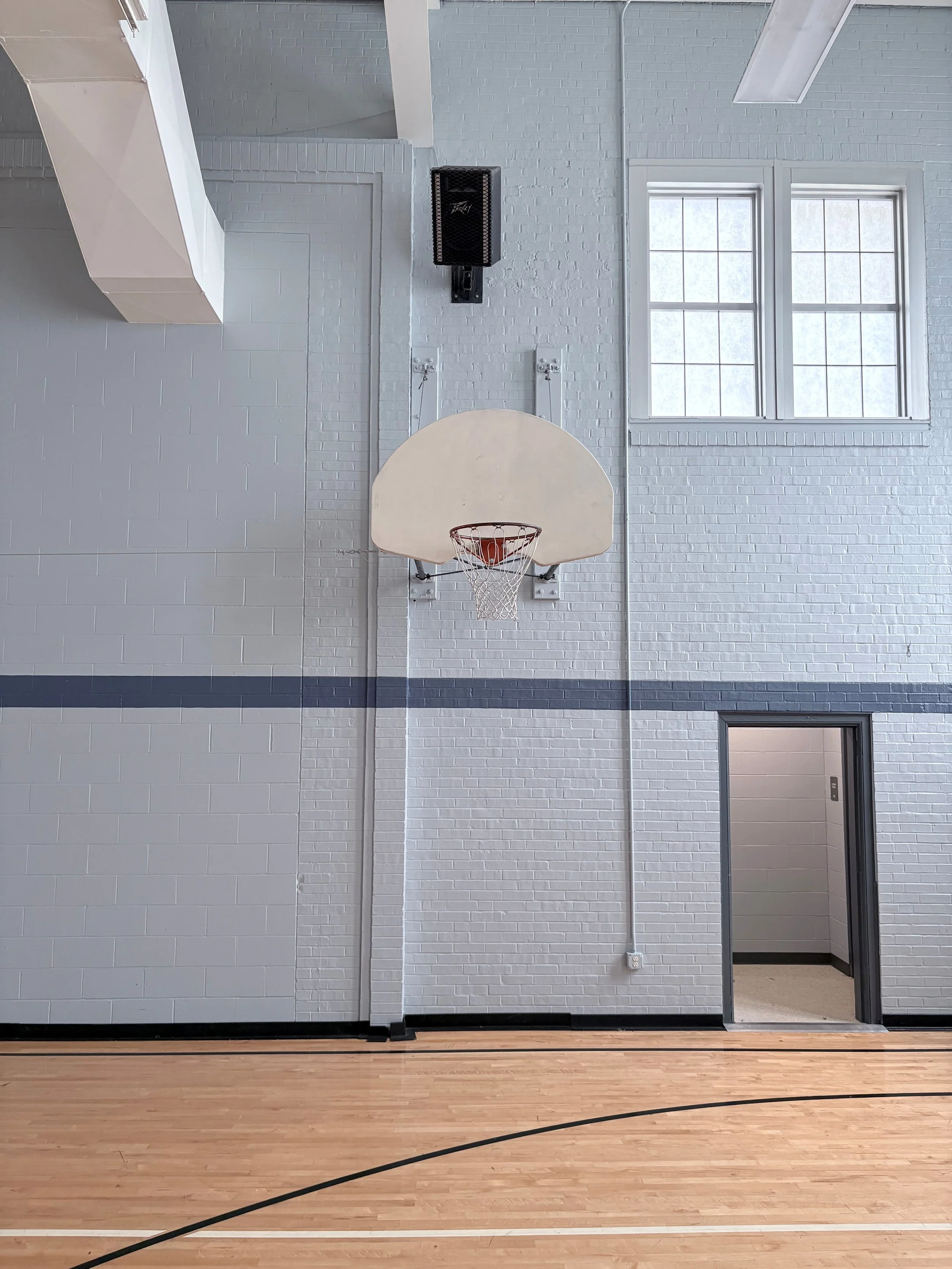 Indoor gymnasium with a basketball hoop mounted on a white brick wall, large windows, and a door on the right side leading to another room.