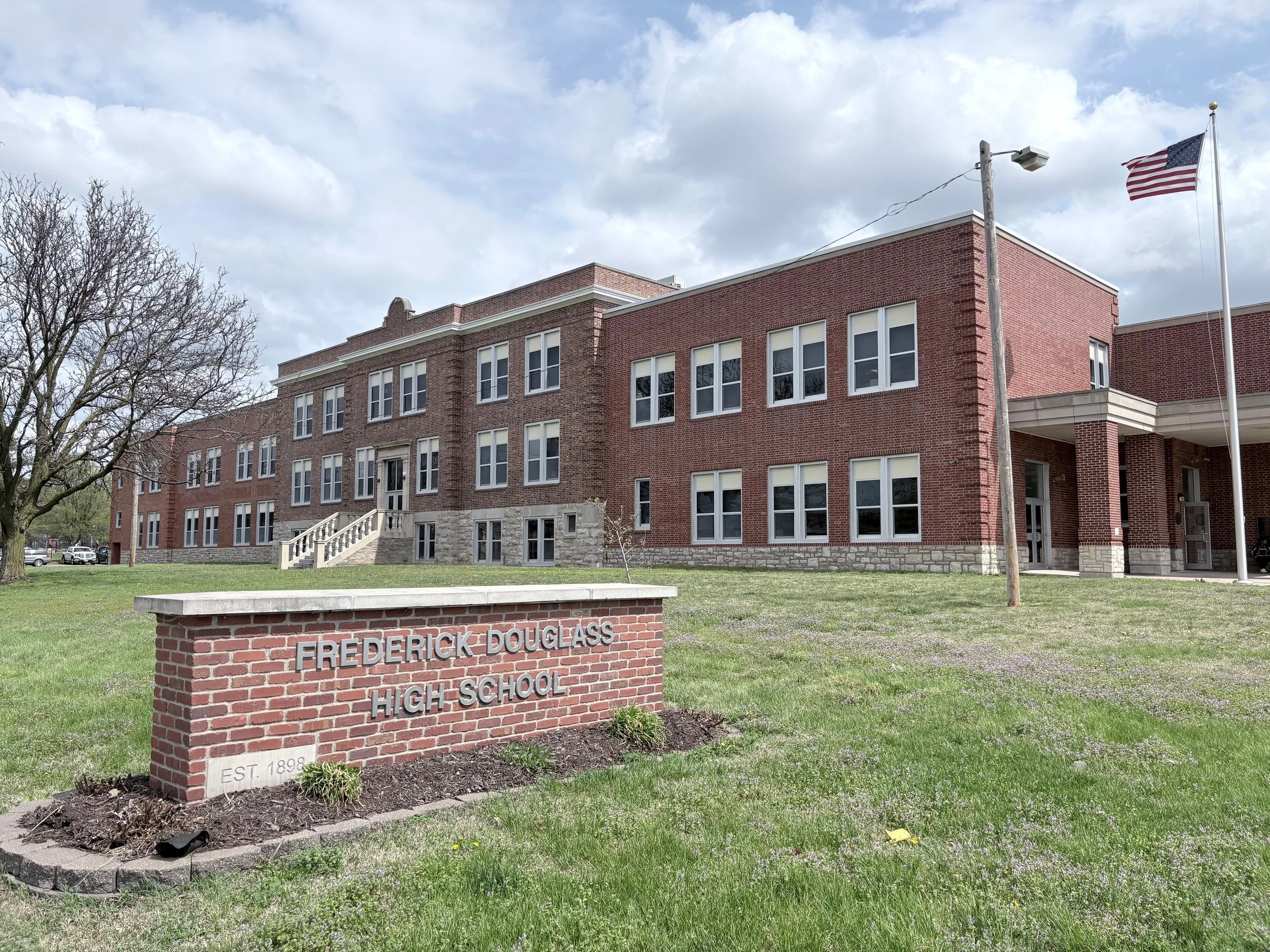 Brick building with multiple windows, a flagpole with an American flag, and a brick sign reading 'Frederick Douglass High School' in front.