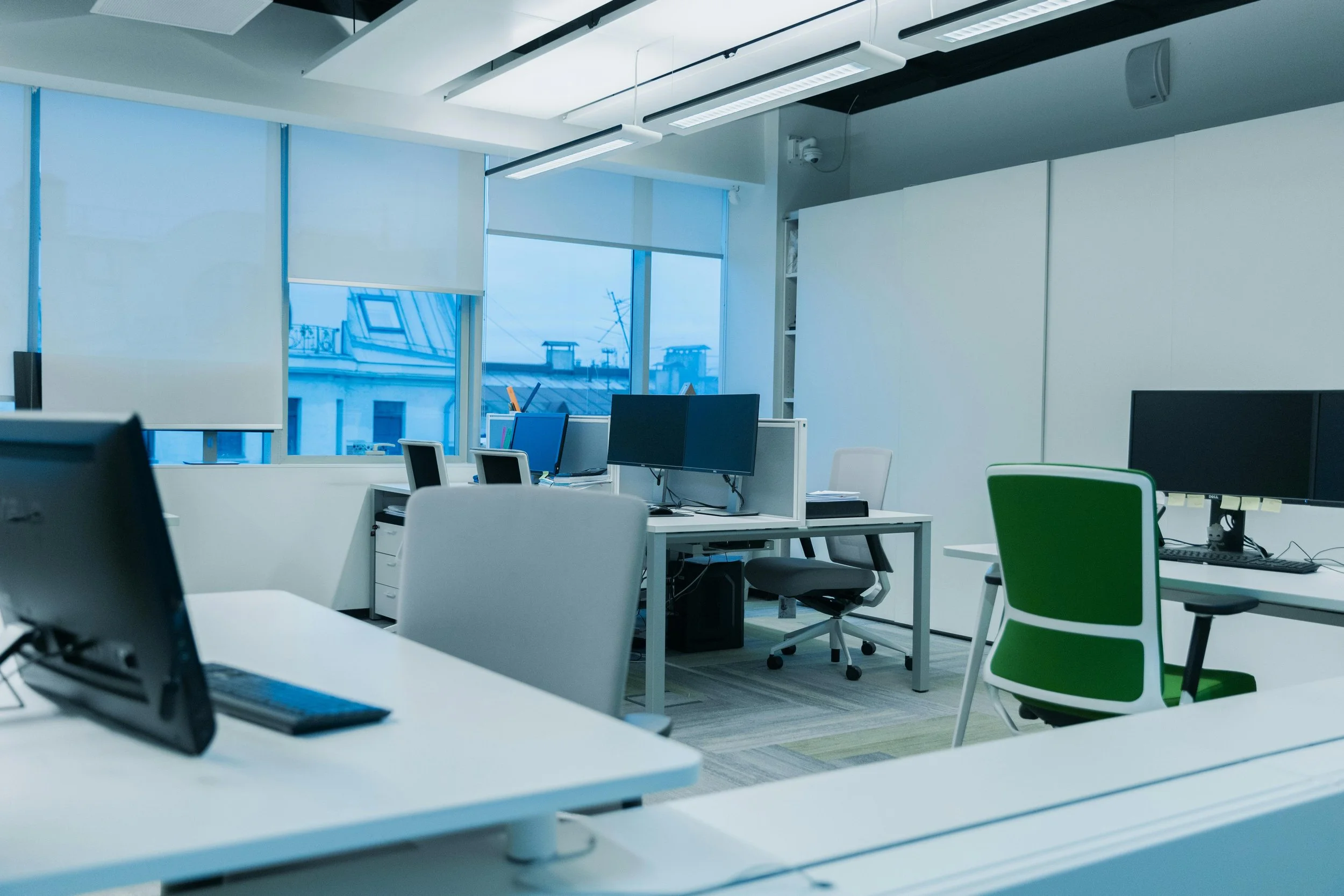 An office space with white desks, chairs, and computer monitors in front of large windows with blinds. The view outside shows rooftops and utility poles.