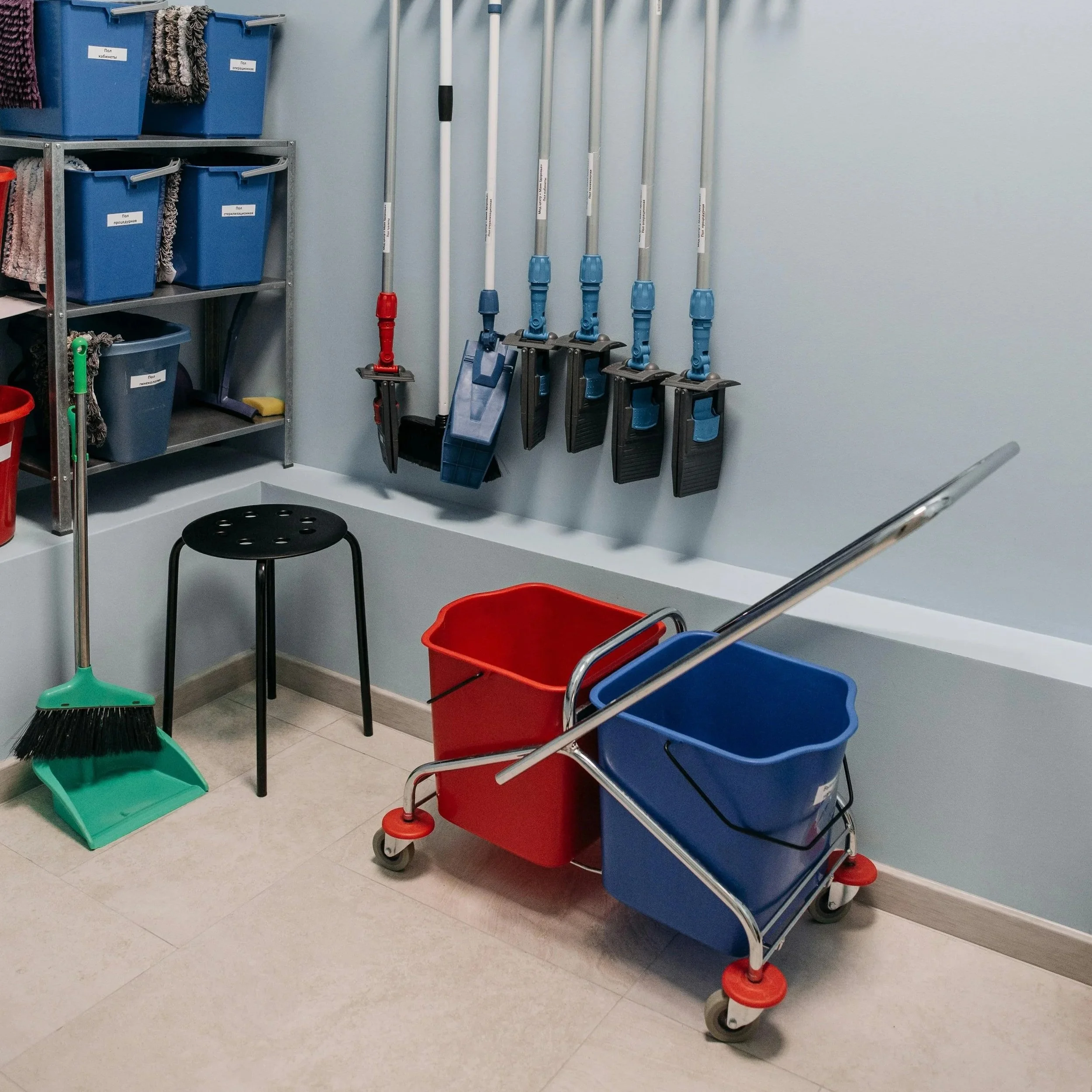 A cleaning cart with blue and red bins and cleaning tools such as mops, brooms, and dustpans in a janitorial closet or cleaning supply storage area.