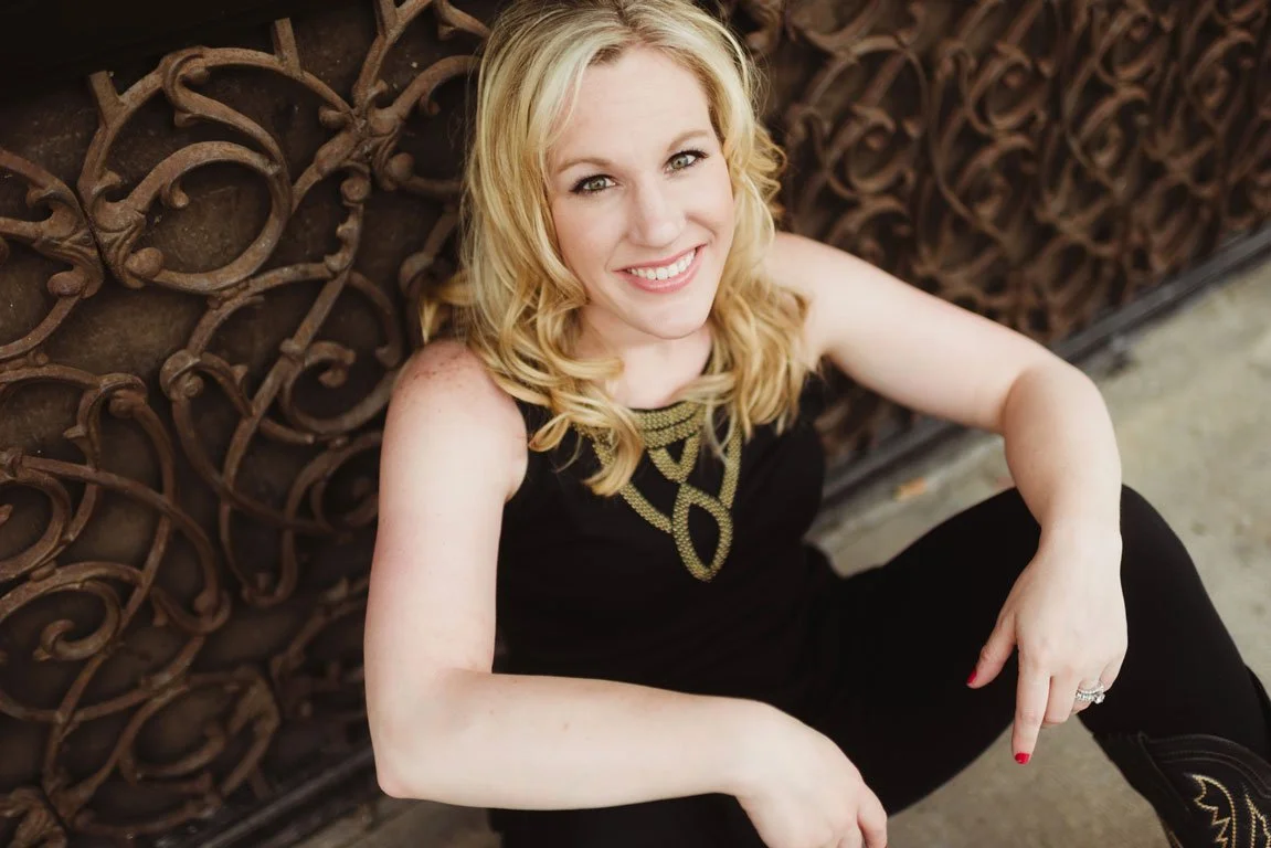 A smiling woman with blonde hair, wearing a black top with gold embellishments, sitting on the ground next to a decorative metal wall.