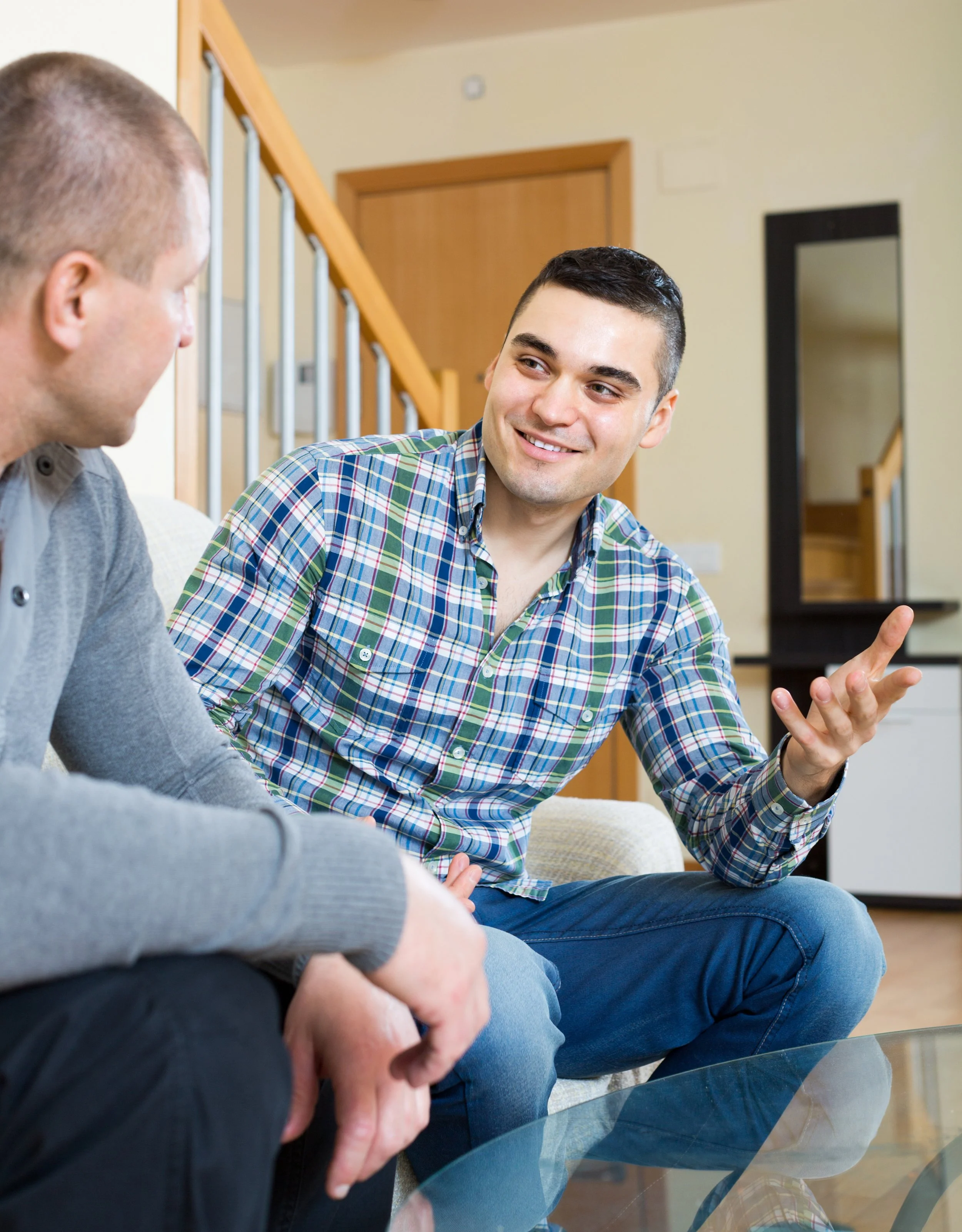Two men sitting on a sofa, engaged in conversation. The man on the right is smiling and gesturing with his hand, while the man on the left turns towards him, listening. A staircase is visible in the background.