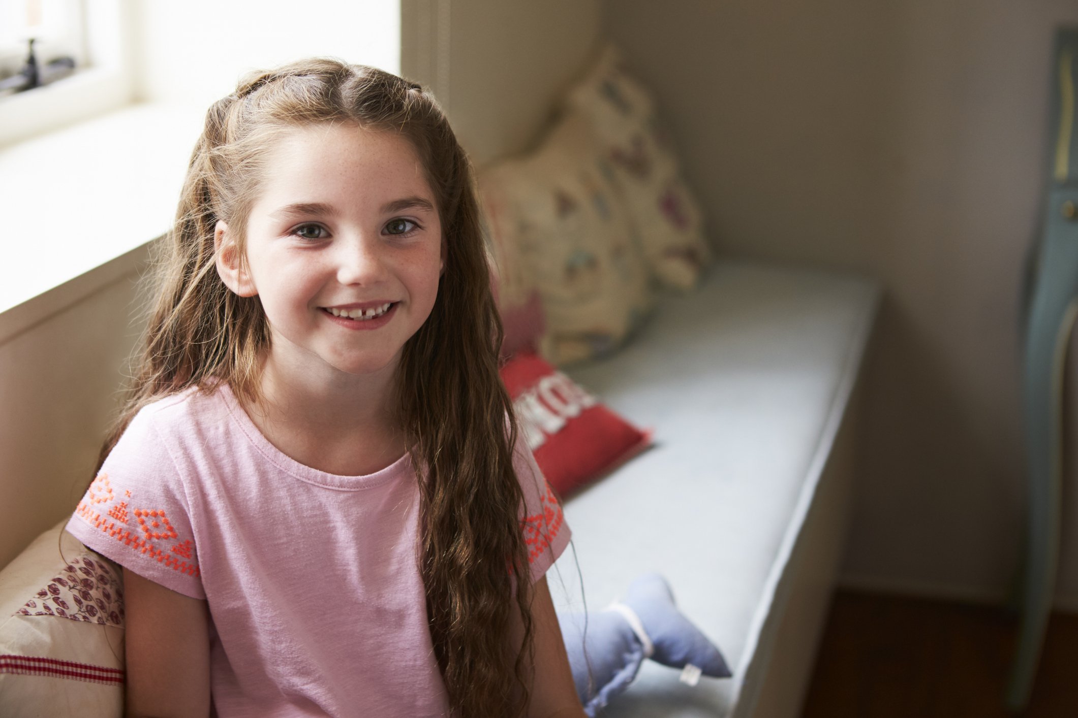A young girl participating in Georgia Autism Spectrum Disorder (ASD) Crisis Homes with long wavy hair and freckles smiling indoors, sitting on a bed with colorful pillows.
