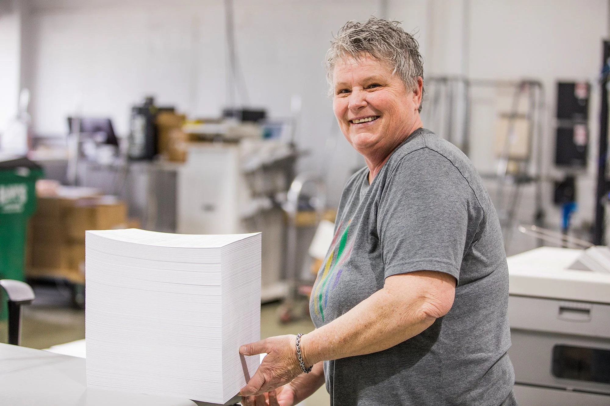 A Tandem Solutions worker with short gray hair handling a large stack of white paper in an industrial or office setting.