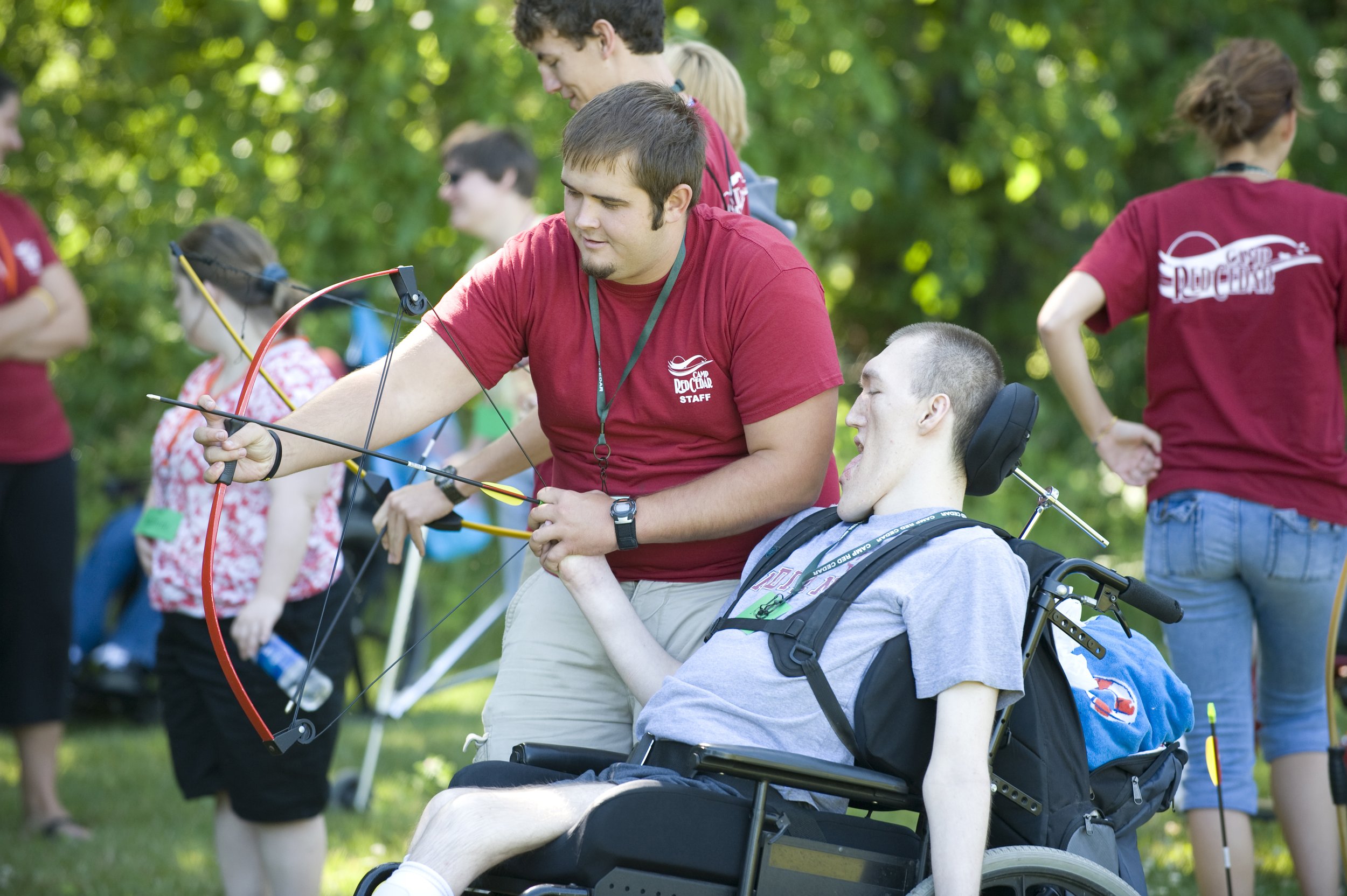Image of a Camp Red Cedar staff worker helping a disabled camper in a wheelchair learn how to shoot a bow and arrow.