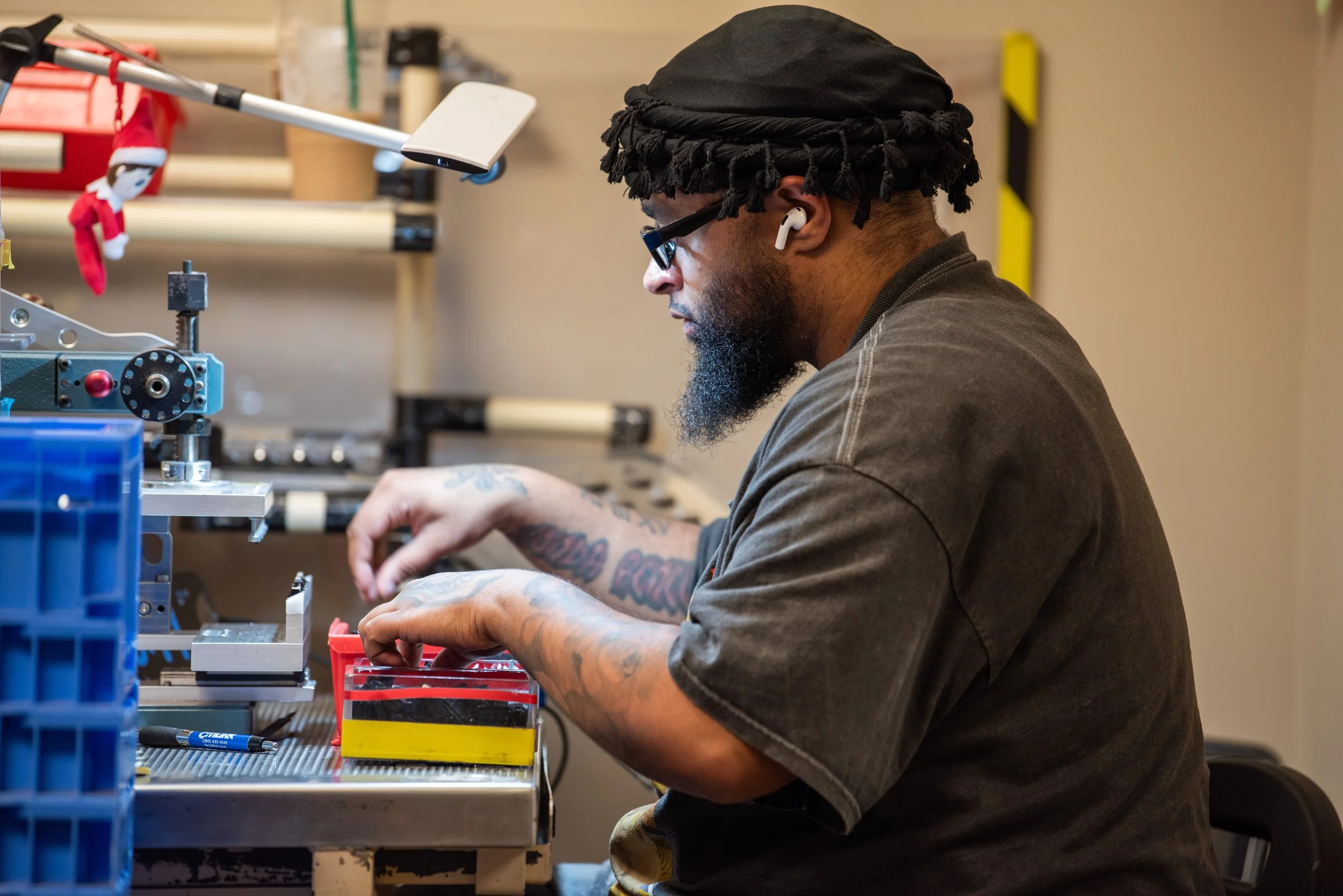 Man working at a workstation, wearing glasses and wireless earphones, with equipment and organized tools around.