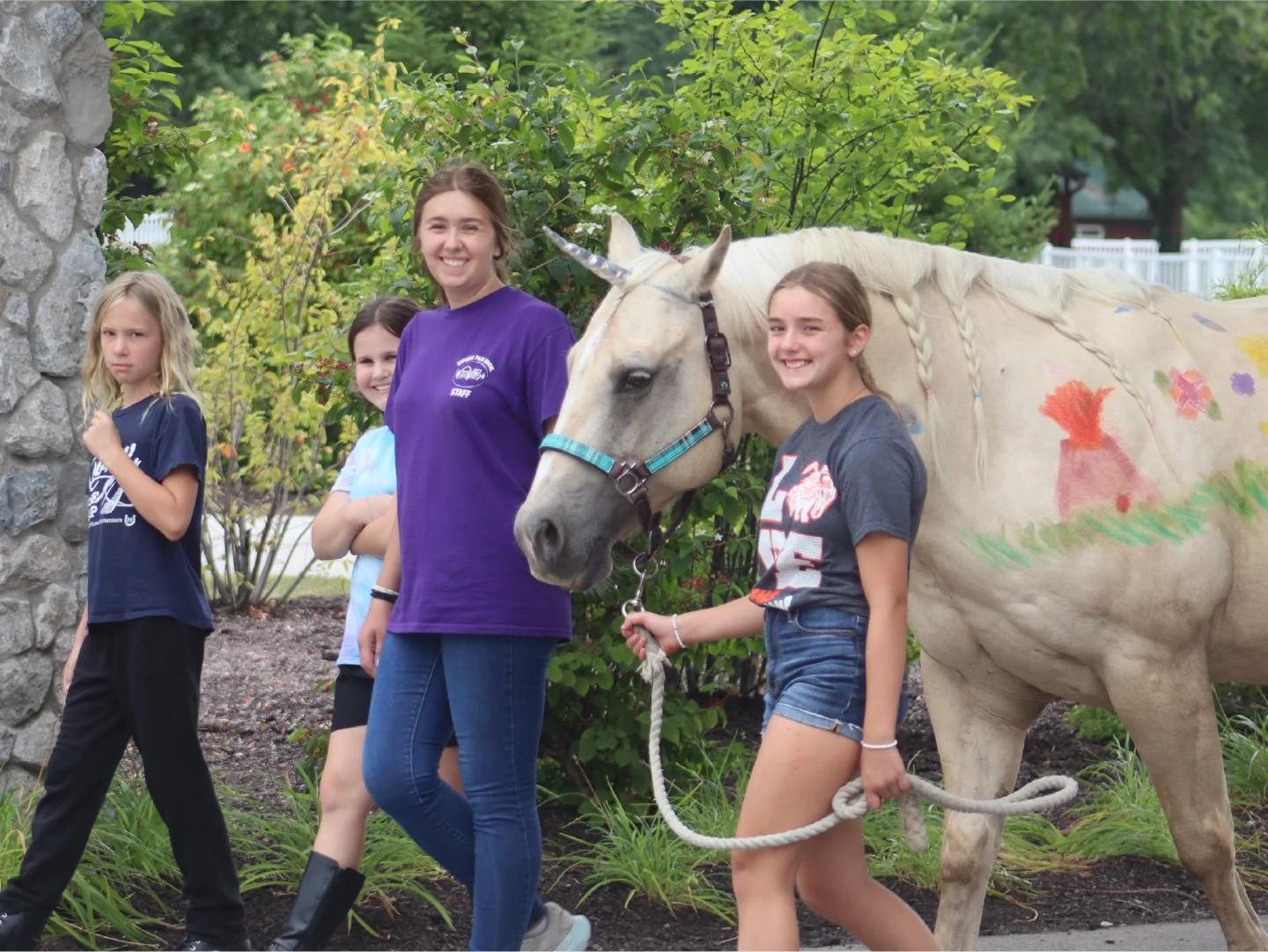 Camp Red Cedar summer campers with a painted white horse, one girl holding the horse's lead rope, green trees and shrubs in the background.
