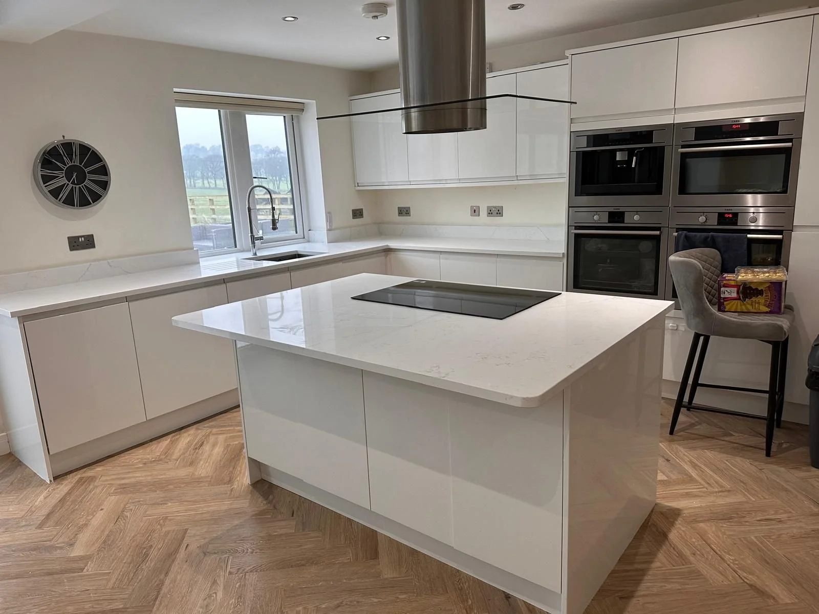 Modern kitchen with white cabinets, a marble countertop island, a window above the sink, stainless steel appliances, and a chair with a box of food on it.