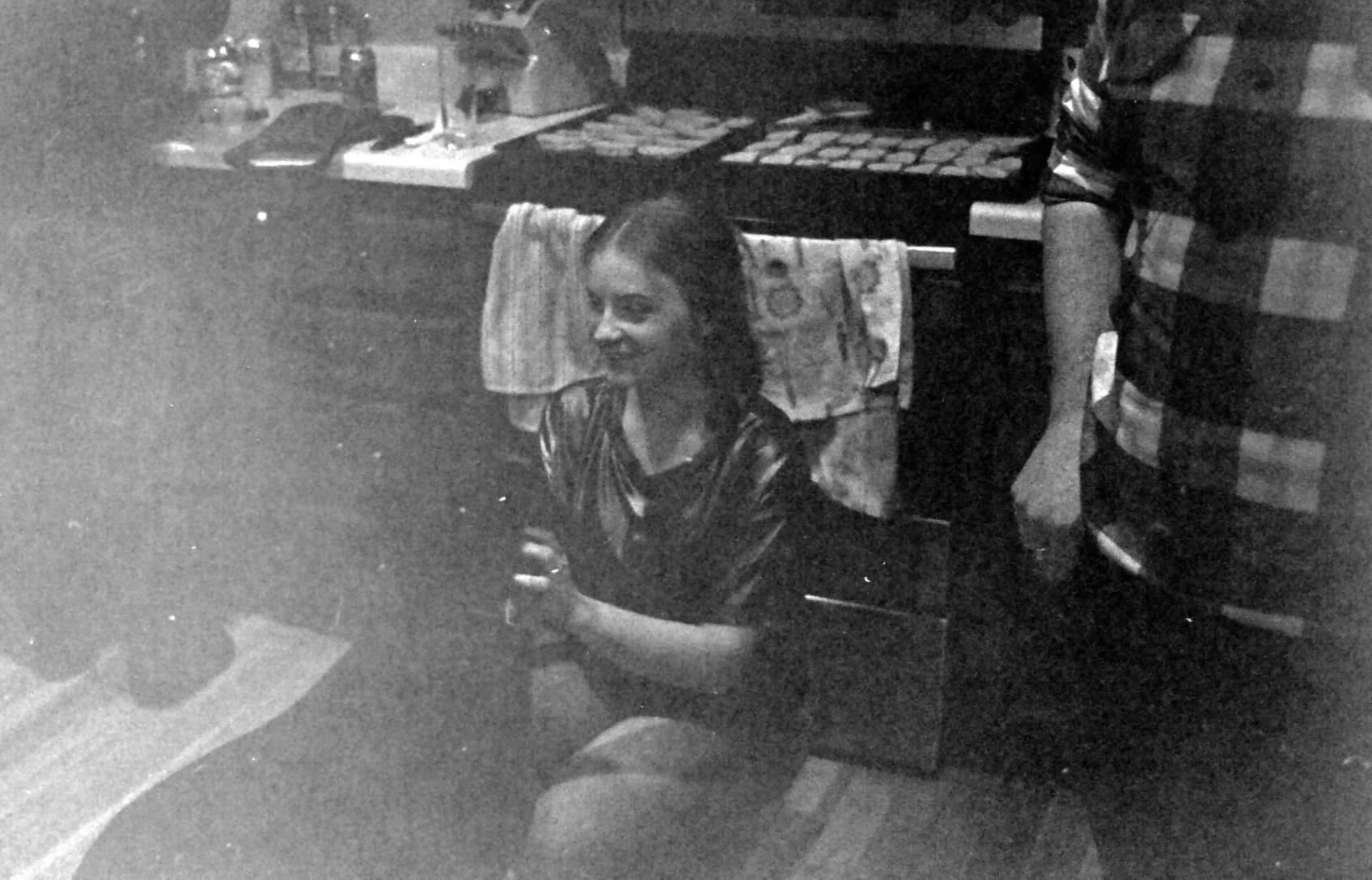 A woman with dark hair sitting on a chair, smiling, in a room with shelves, towels, and various objects in the background.