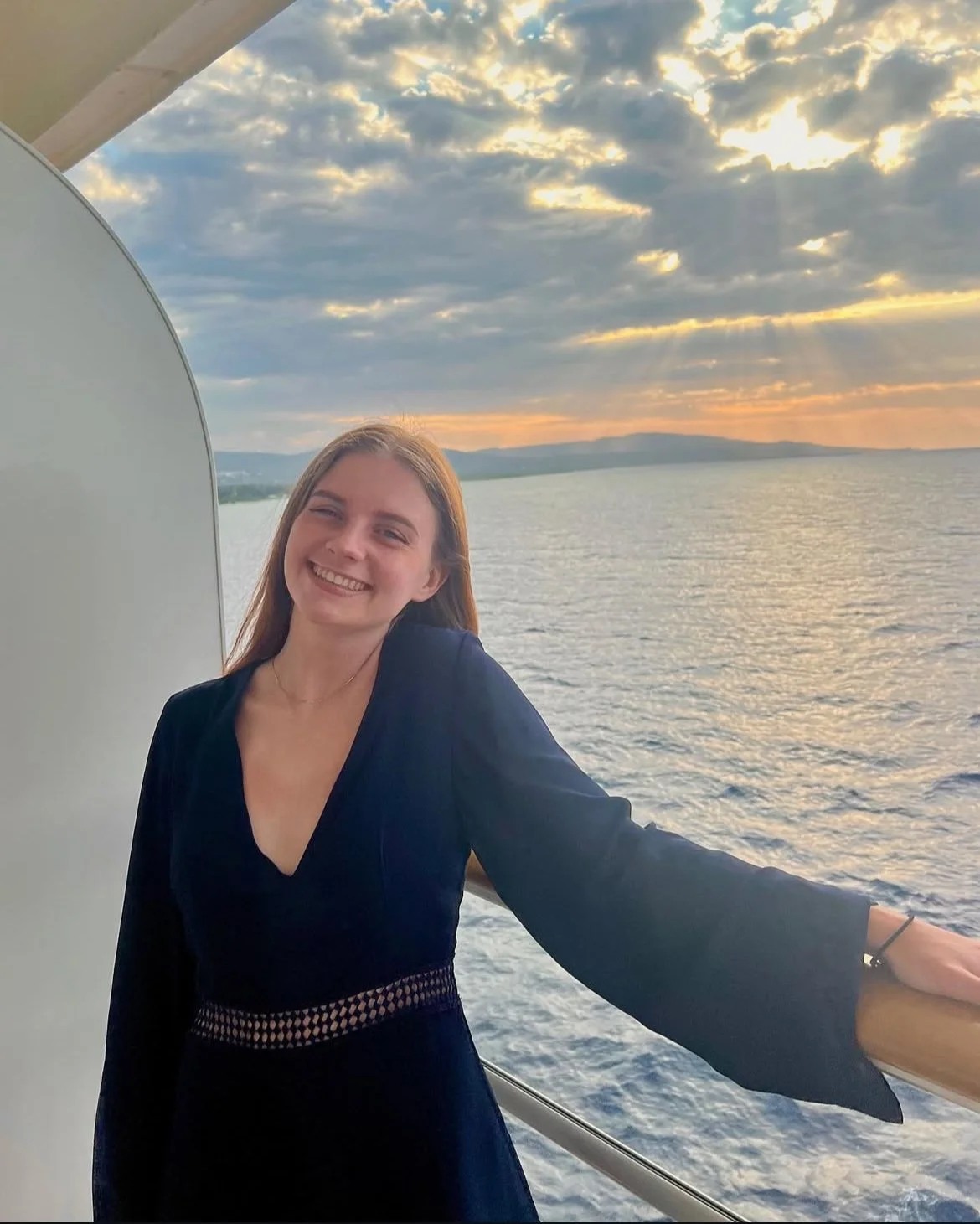 A young woman smiling and standing on a boat with a scenic view of water and a partly cloudy sunset sky in the background.