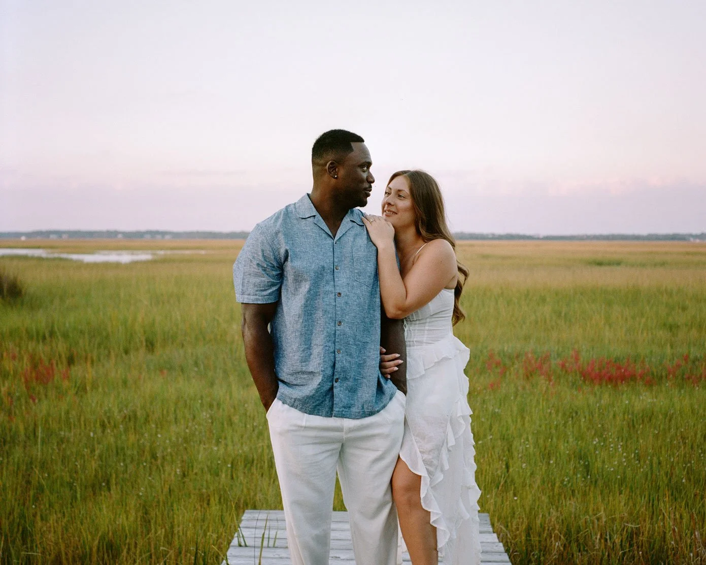 A couple standing on a wooden pathway in a marsh, gazing at each other, with a pastel-colored sky in the background.