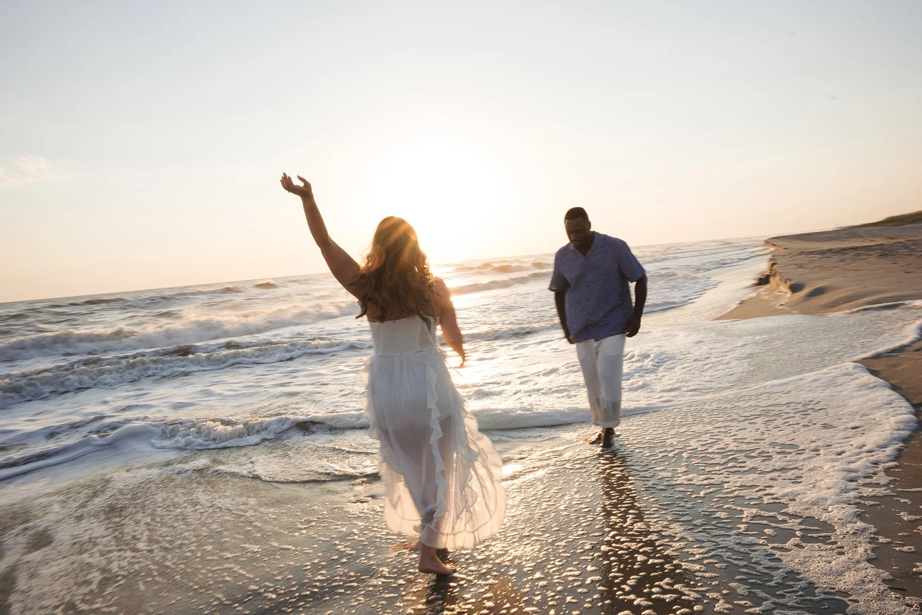 A woman in a white dress and a man in a blue shirt and white pants walking on the beach during sunset with waves in the background.