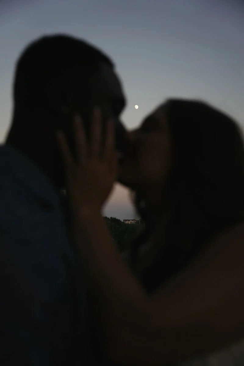 Silhouette of a couple kissing outdoors at dusk with the moon in the background.
