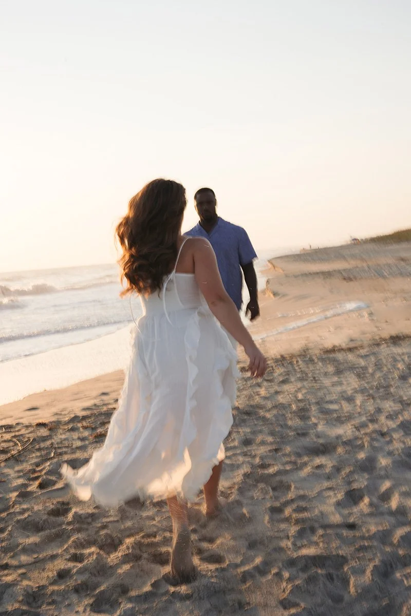 A woman in a white dress walking on the beach with a man in a blue shirt in the background during sunset.