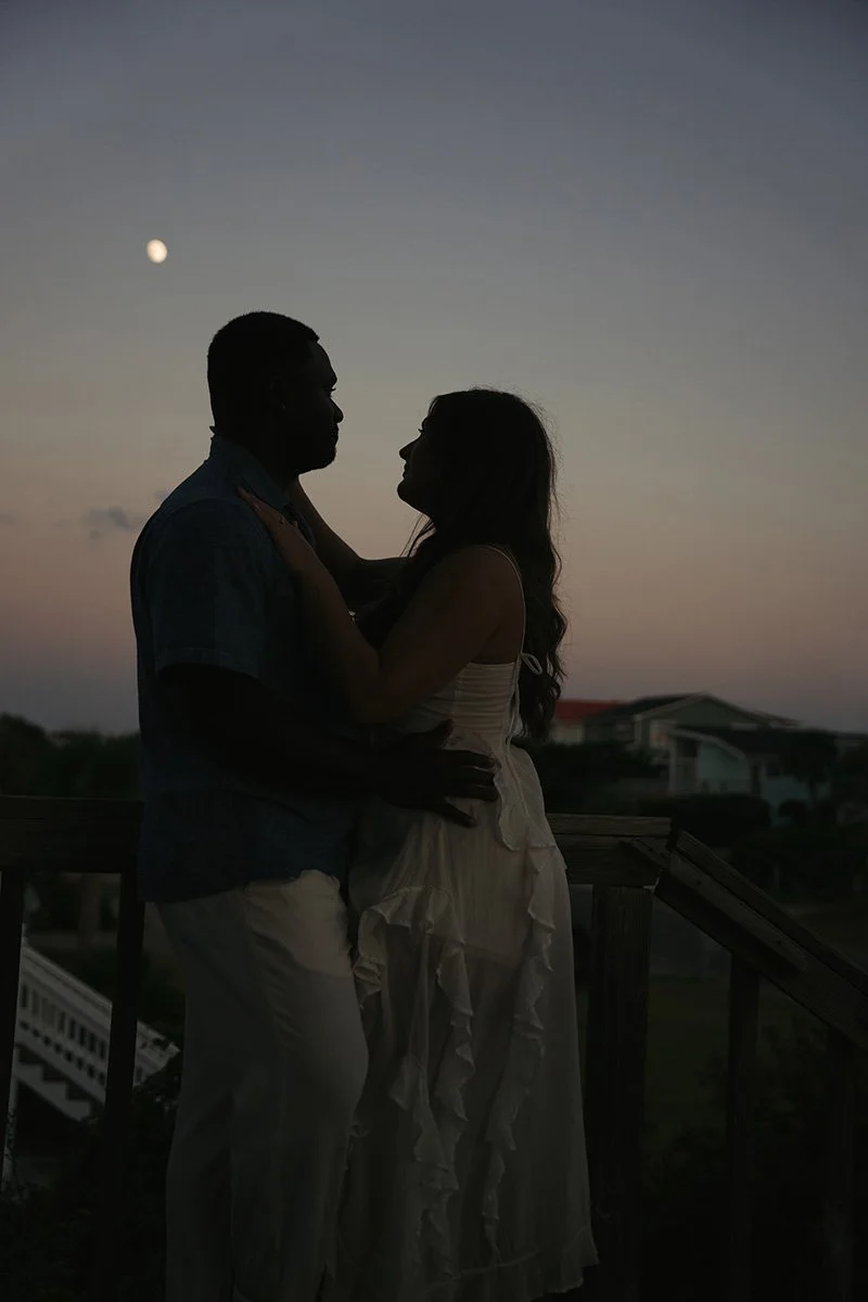 Silhouetted couple embracing during sunset with a visible moon in the sky and houses in the background.