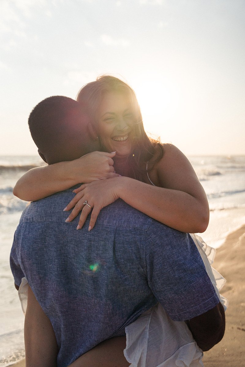 A woman with long hair, smiling, hugging a man on the beach during sunset, with the ocean in the background.