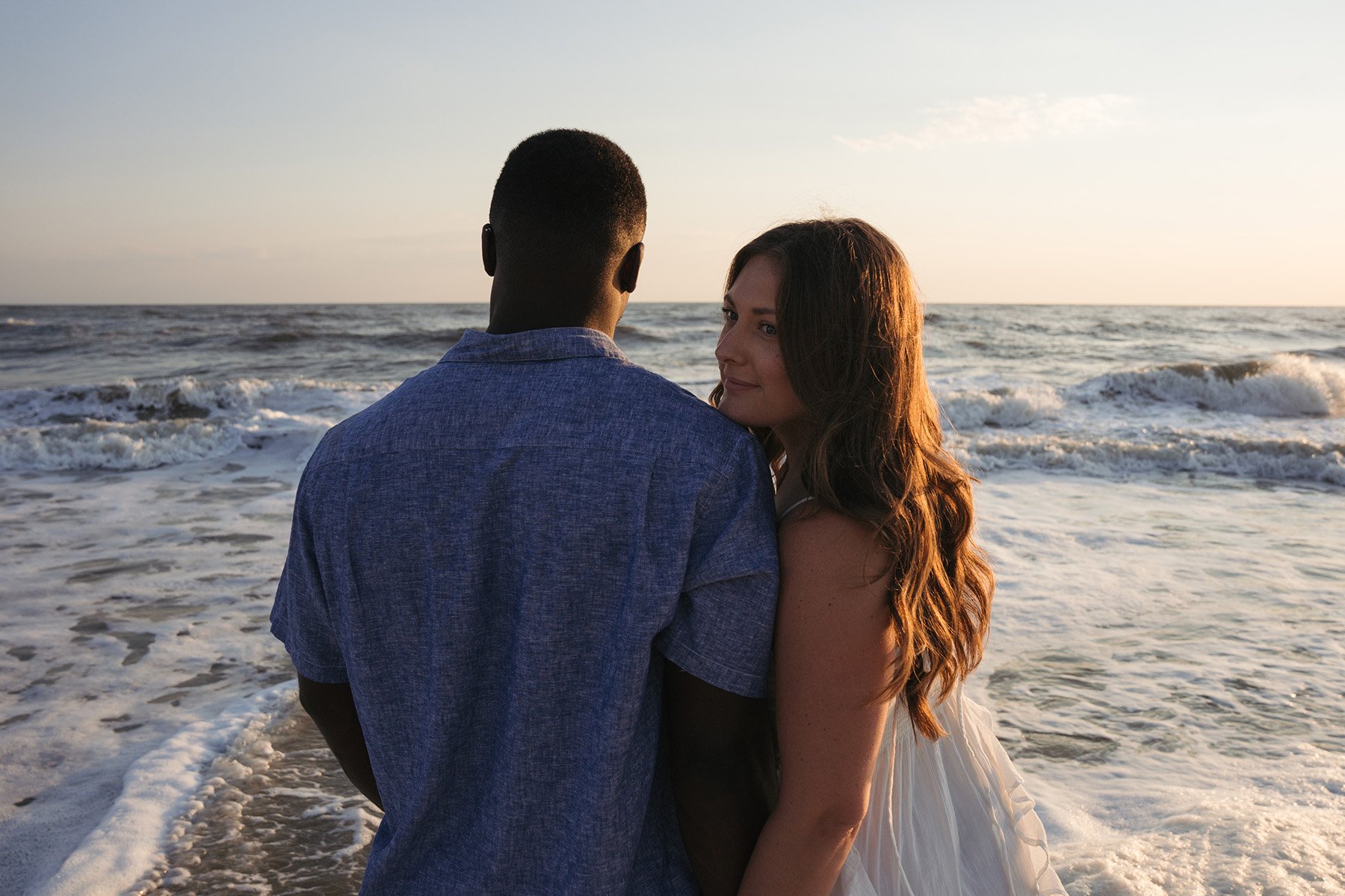 A man and woman stand close together on the beach during sunset, looking at each other with the ocean waves behind them.