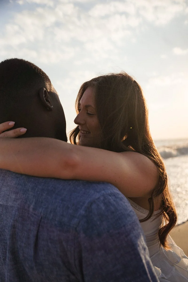 A couple embracing on the beach during sunset, with the woman smiling and holding the man's face, the ocean in the background.