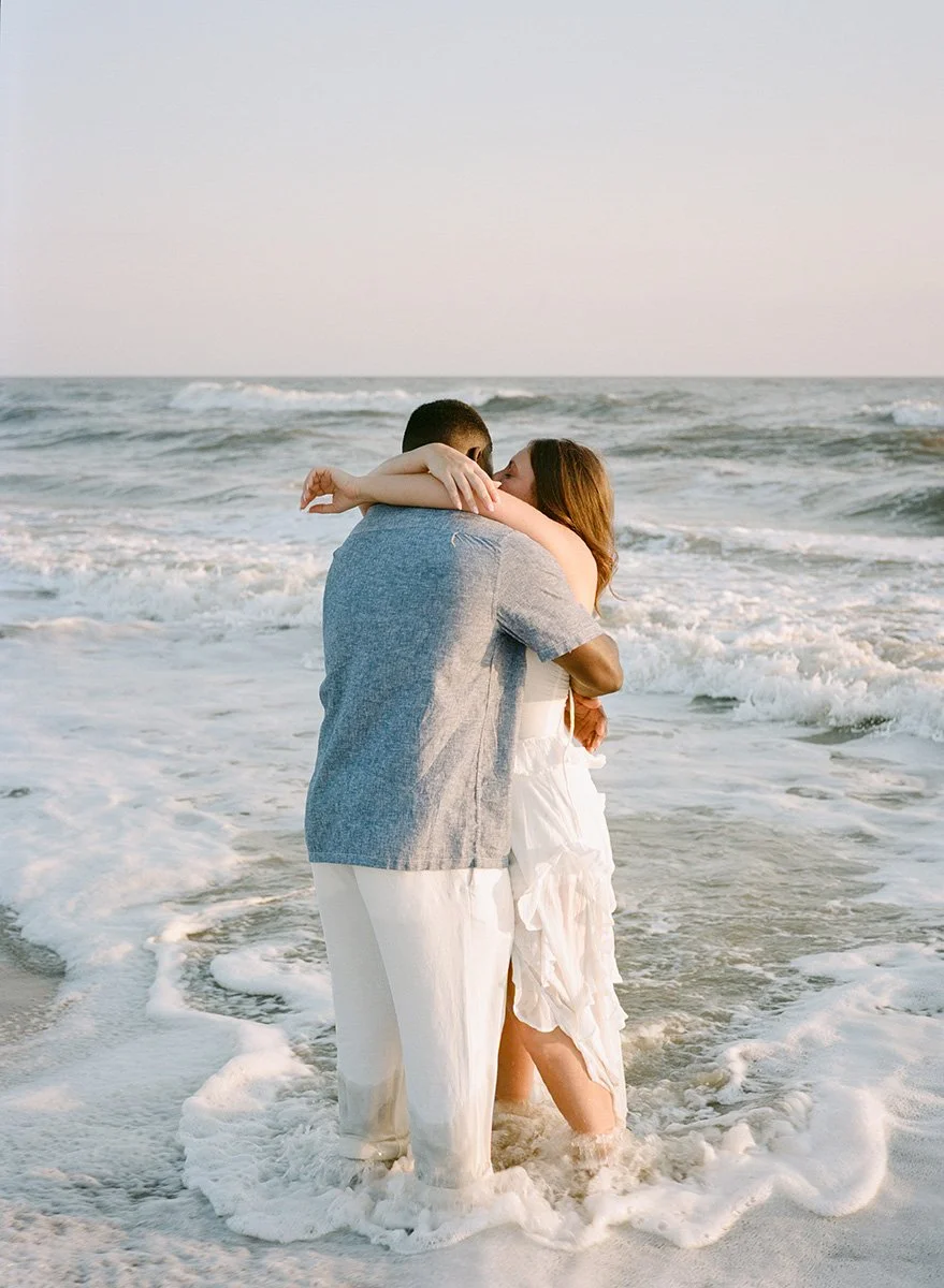 A couple hugging in the ocean at sunset, with waves around their legs.