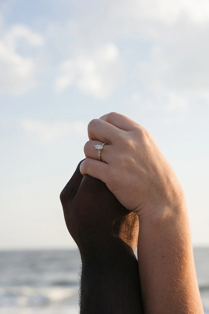 Two hands, one light-skinned and one dark-skinned, holding each other with a sky and ocean in the background. The light-skinned hand has an engagement ring with a large diamond.